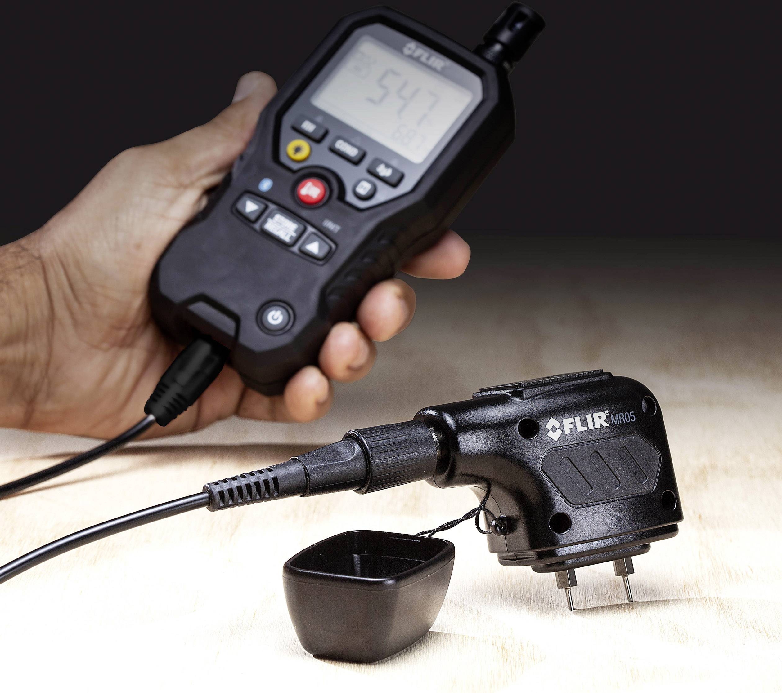 Urządzenie ręczne wyświetla wartość '54.1'. Jest podłączone przewodem do czujnika leżącego na powierzchni drewna.