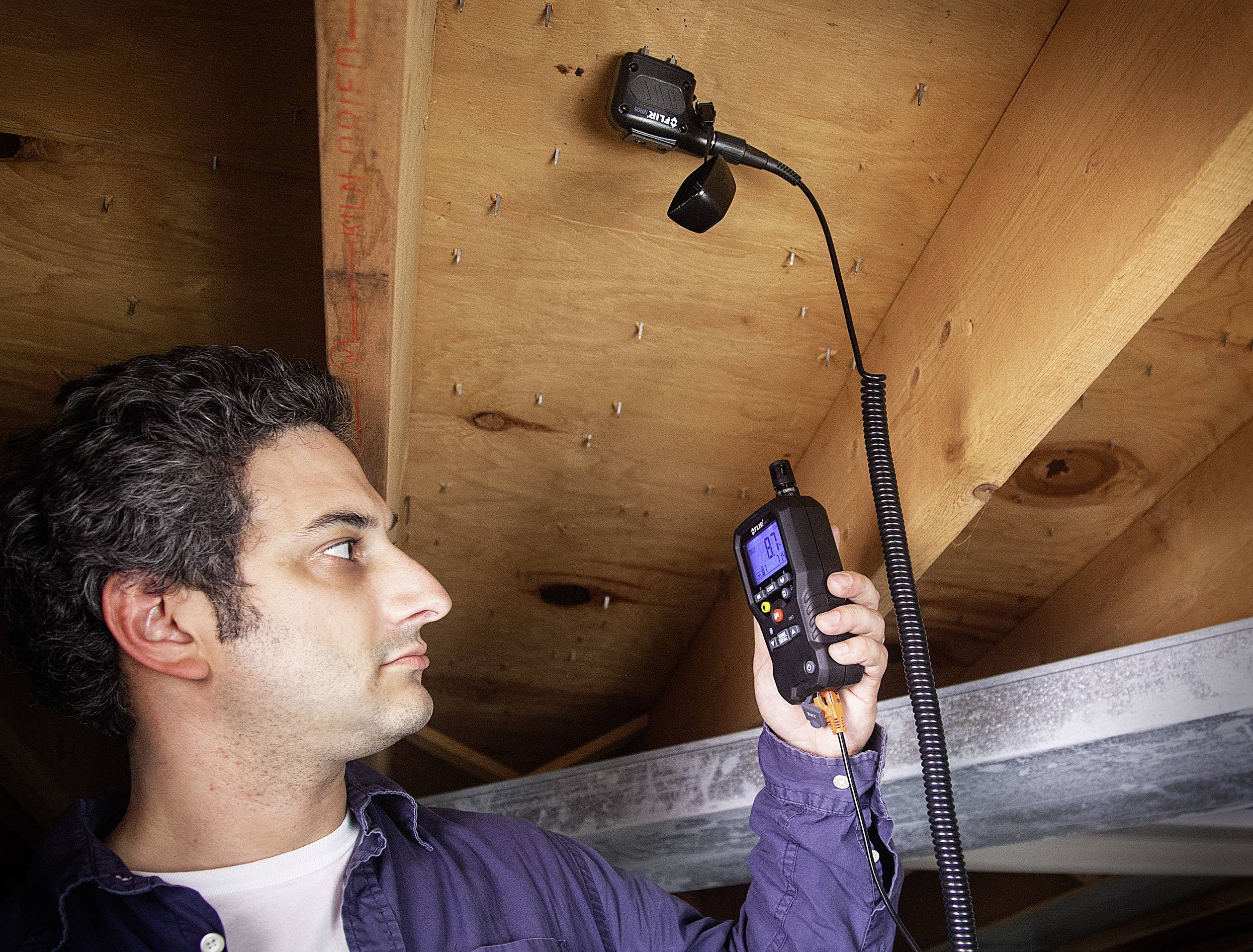 Mężczyzna używa kamery inspekcyjnej do sprawdzenia przestrzeni między belkami drewnianymi. Kamera jest elastyczna i posiada wyświetlacz.