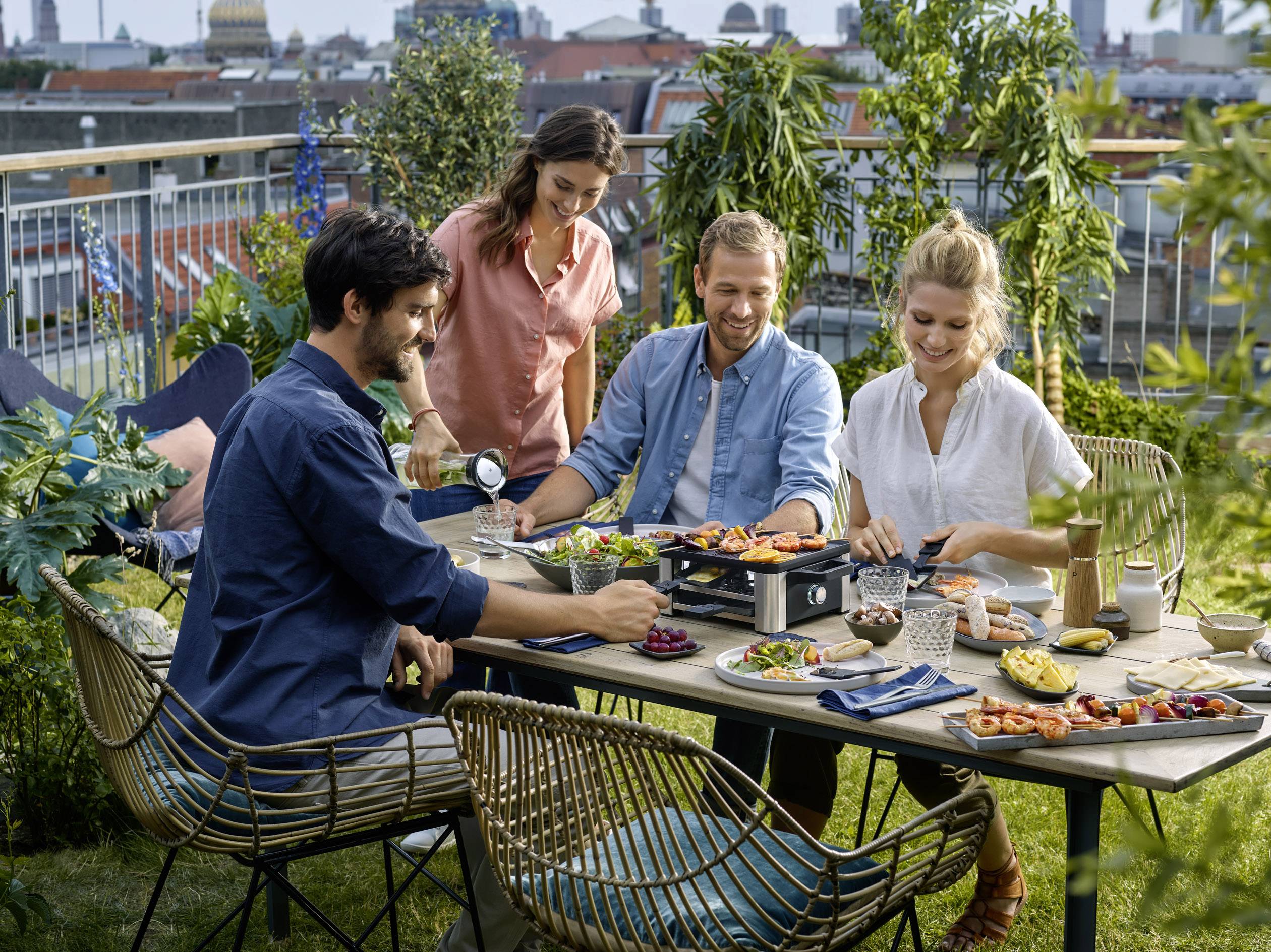 Cztery osoby siedzą na tarasie wokół stołu, grillując i śmiejąc się. W tle widać miejską panoramę miasta.