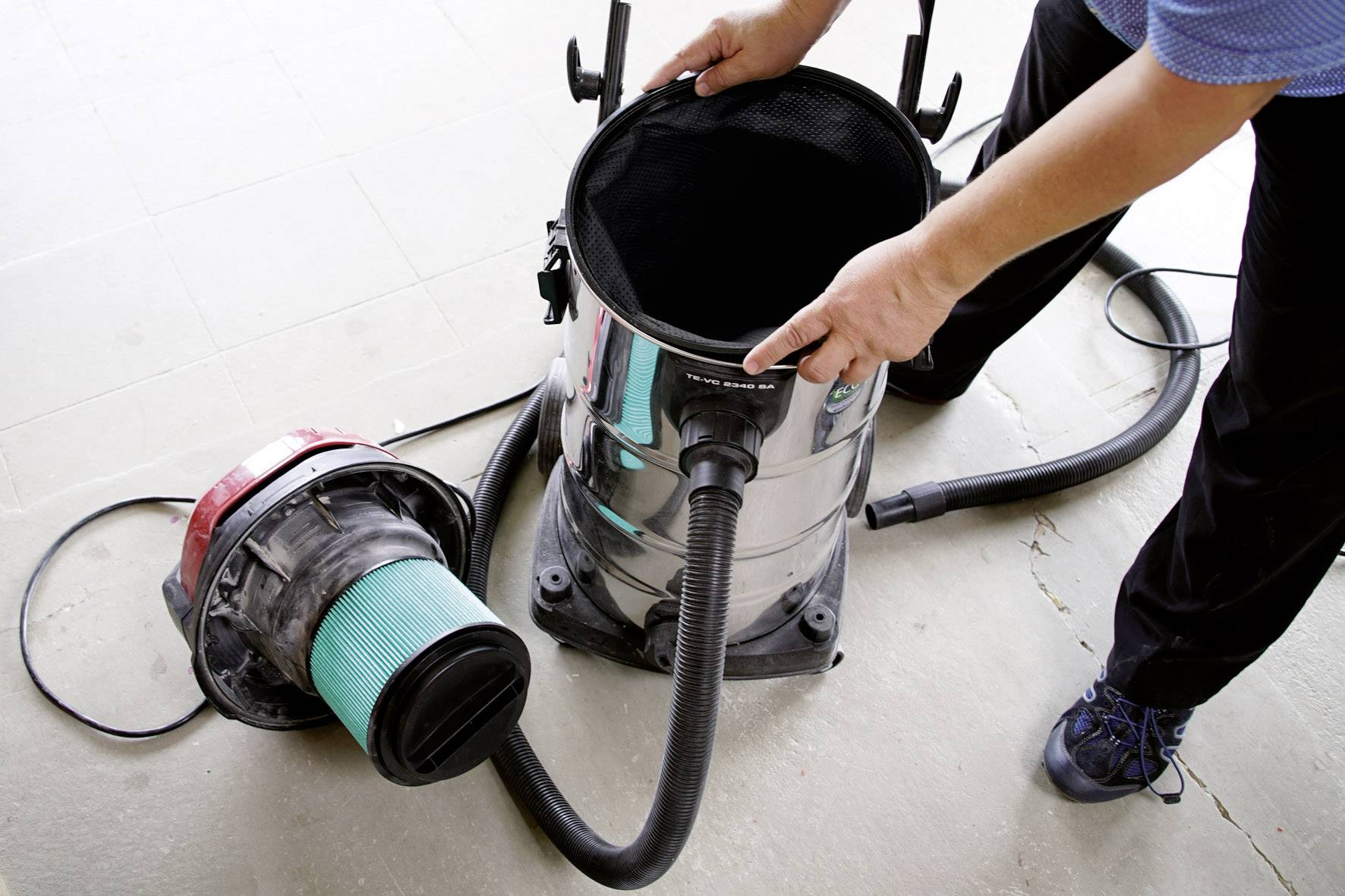 Osoba otwiera pojemnik odkurzacza, aby sprawdzić lub wymienić filtr. Odkurzacz stoi na podłodze.