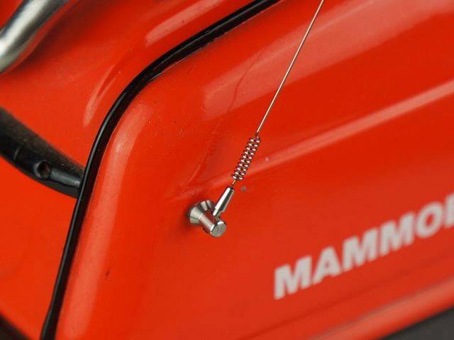 Zbliżenie czerwonego urządzenia z małym metalowym zaworem i przewodem. Napis 'MAMMOT' widoczny jest niewyraźnie w tle.