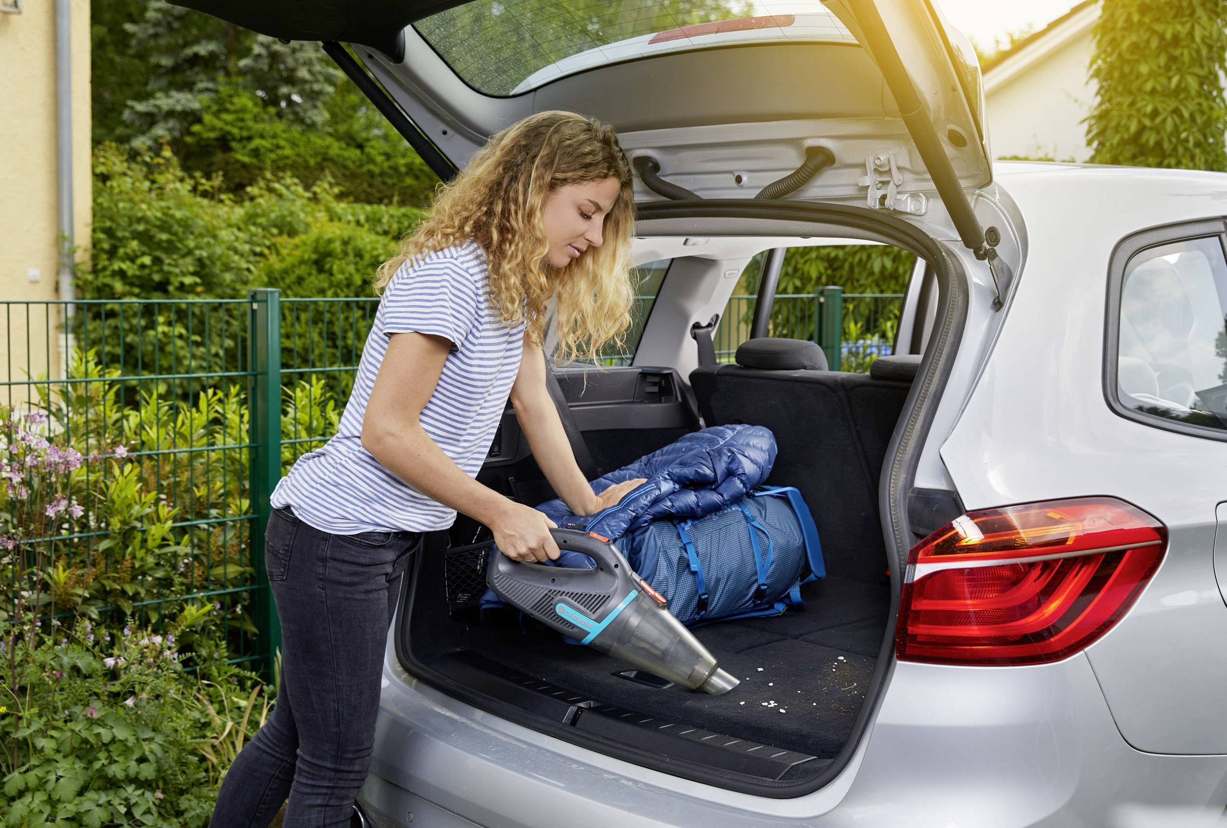 Kobieta czyści bagażnik samochodu przenośnym odkurzaczem ręcznym. W bagażniku znajduje się kurtka i plecak.