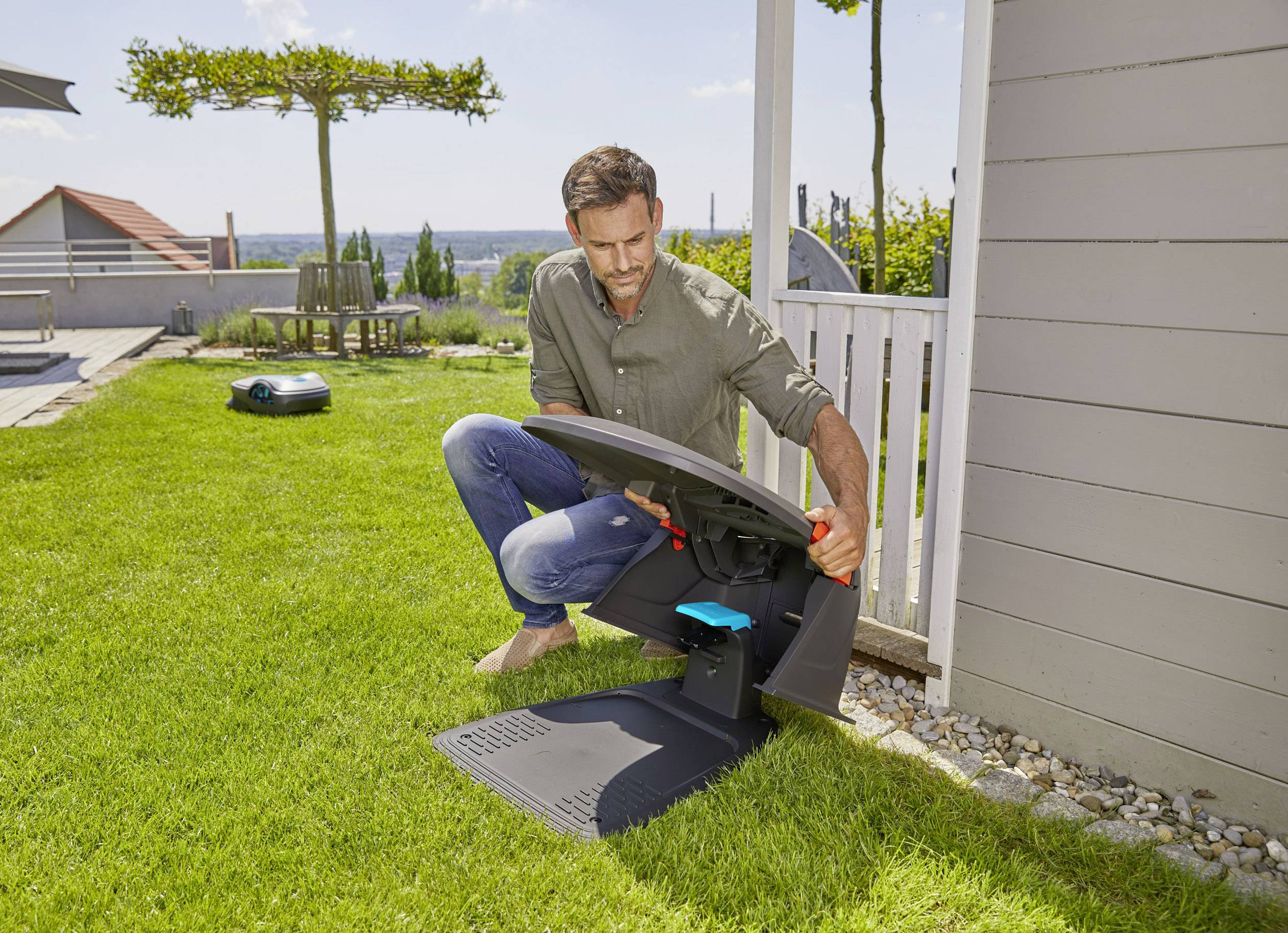 Mężczyzna sprawdza robota koszącego w stacji ładującej w ogrodzie. W tle widoczne są krzesła ogrodowe i drzewa.