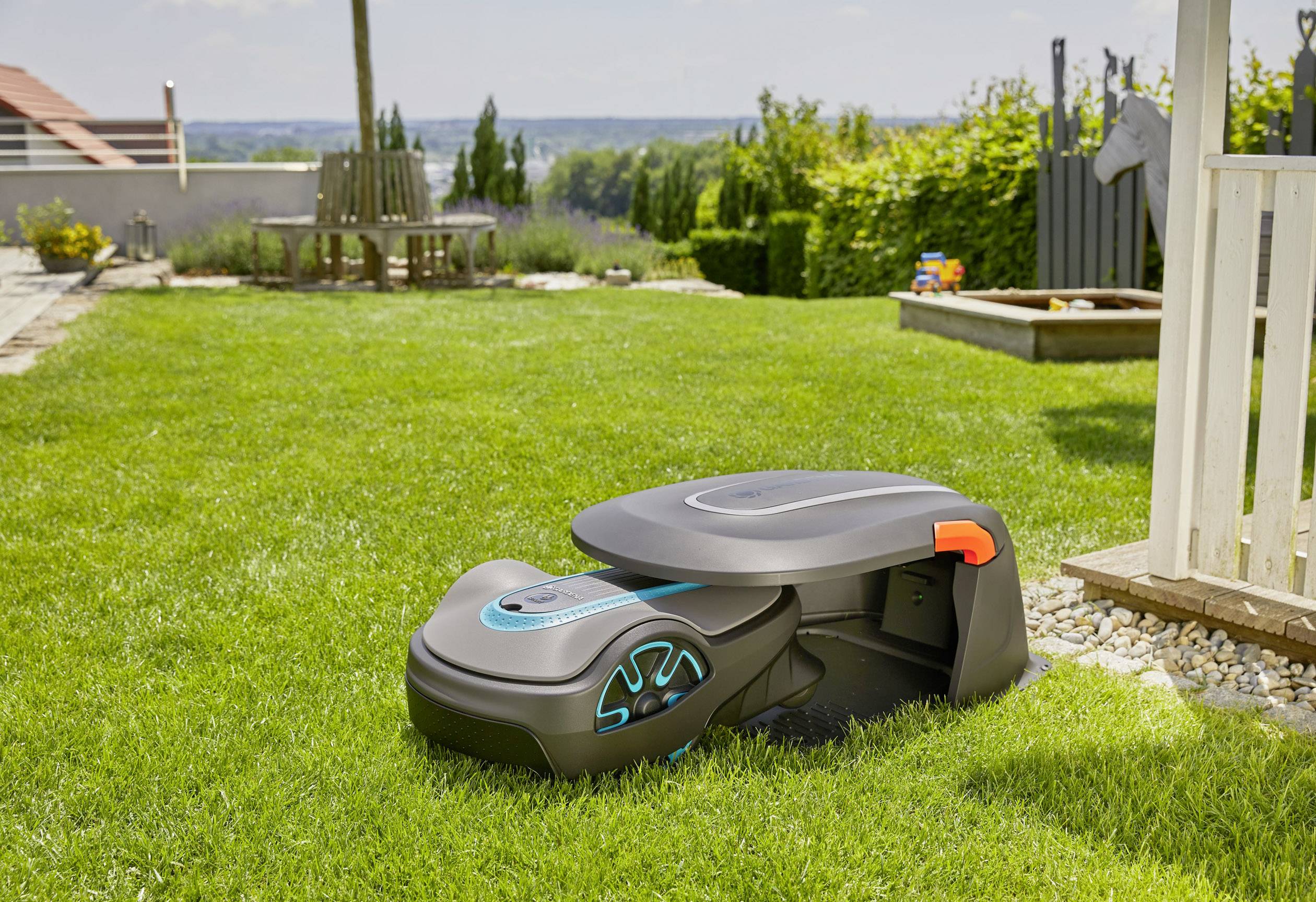 Robotyczny kosiarz na starannie utrzymanym trawniku w ogrodzie, gotowy do pracy. W tle widoczne są drzewa i bezchmurne niebo.