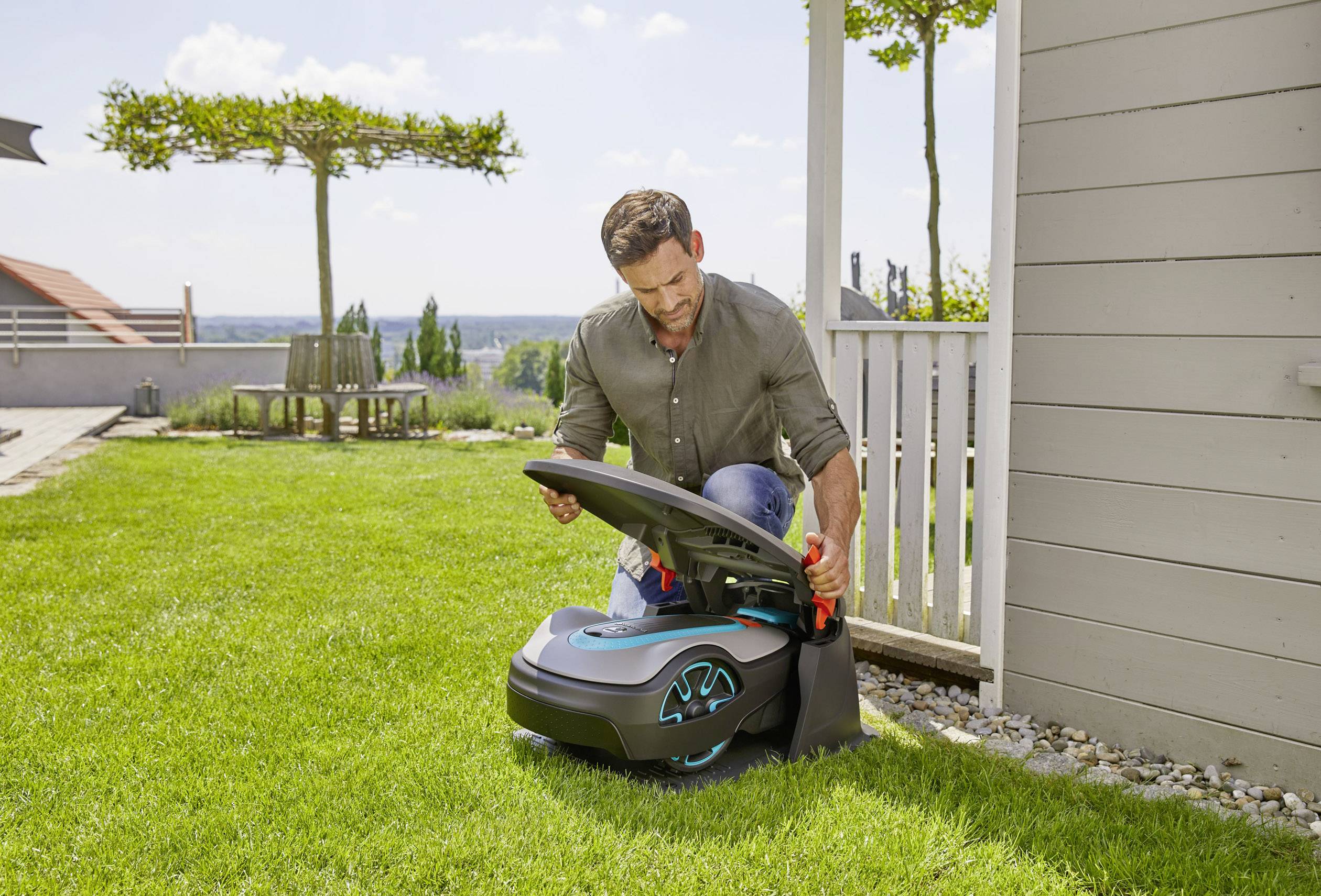 Mężczyzna klęczy na trawniku i sprawdza automatyczną kosiarkę robotyczną obok domu. W tle widać ogród.