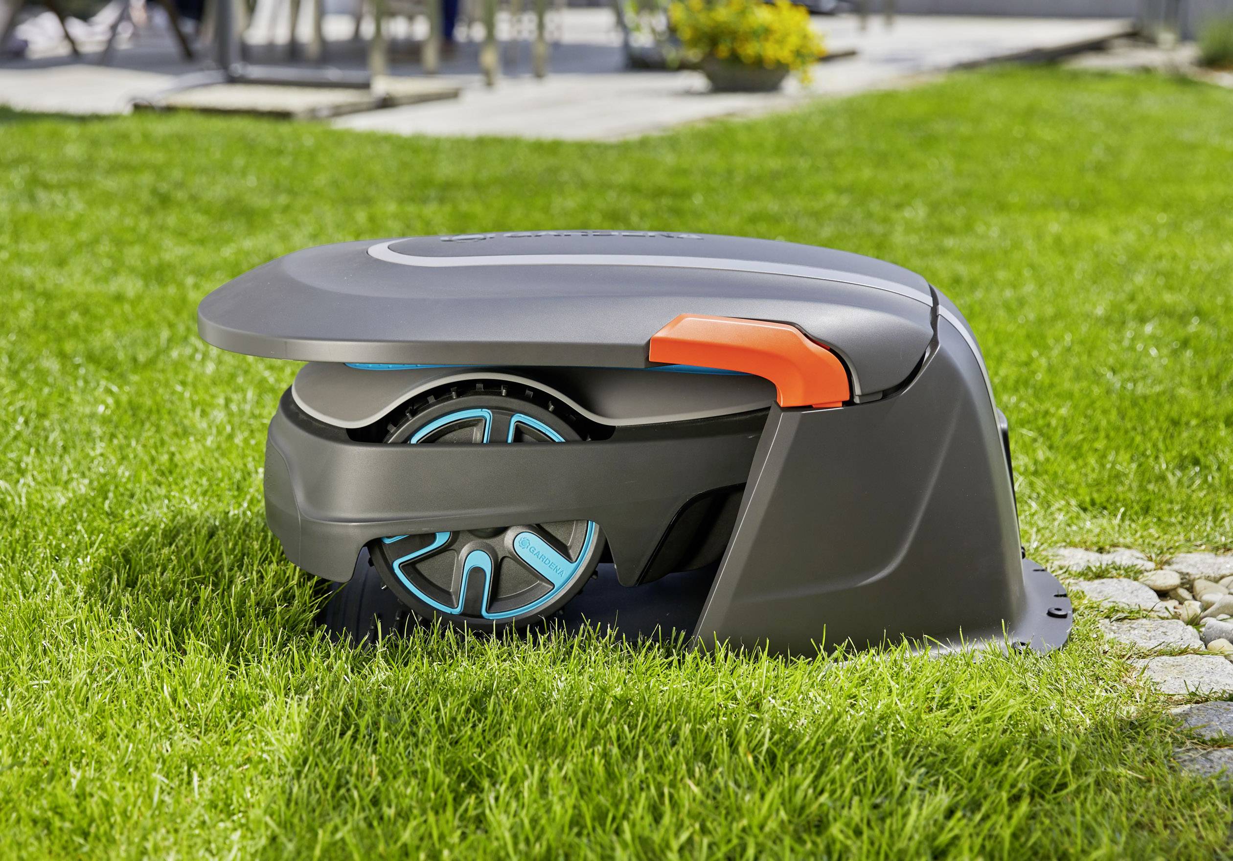 Robot koszący zaparkowany w stacji ładującej na zielonym trawniku na wolnym powietrzu, otoczony kilkoma kwiatami i wybrukowaną ścieżką w tle.