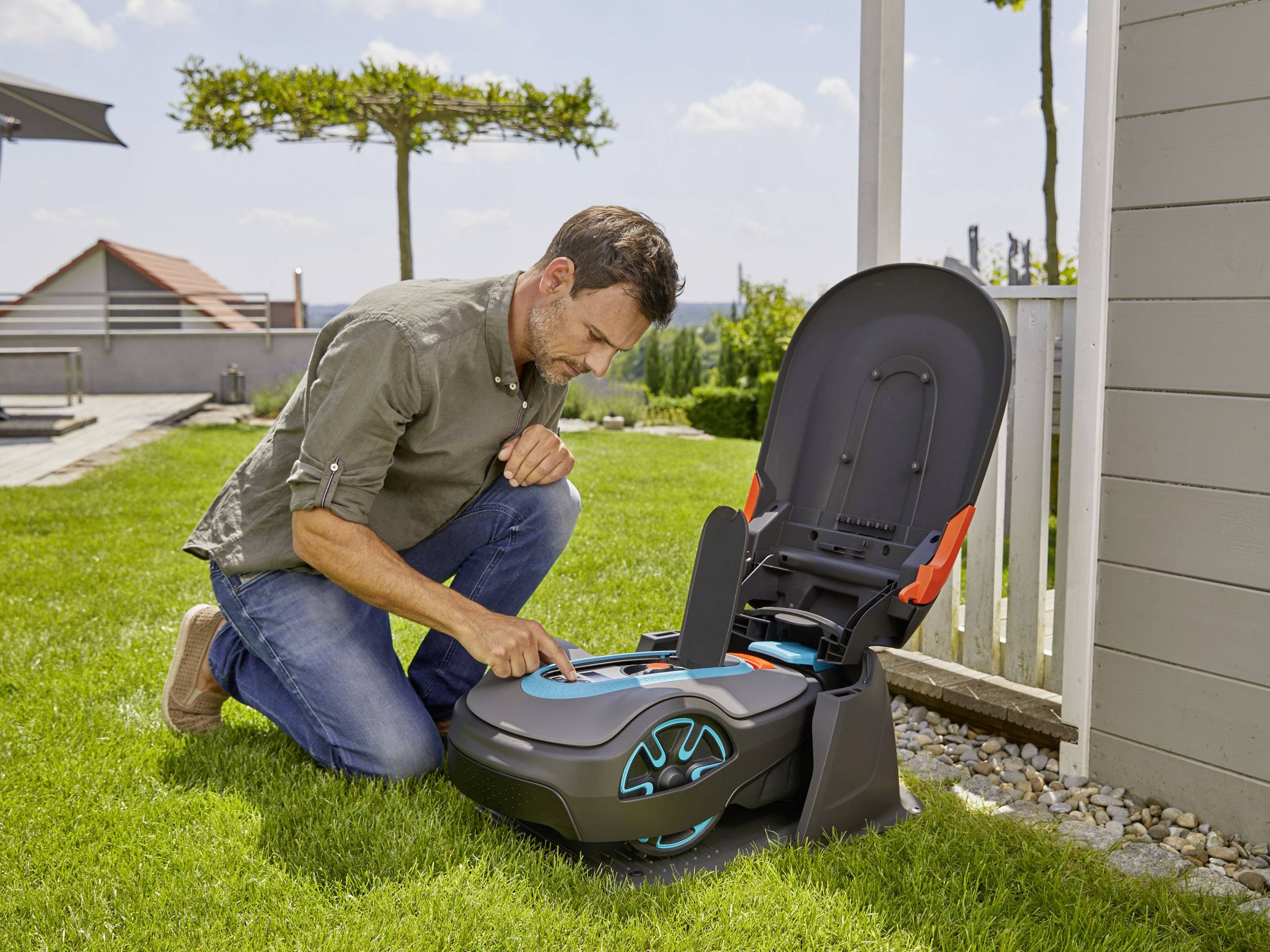 Mężczyzna klęczy w ogrodzie i obsługuje robota koszącego, który stoi w stacji ładującej. W tle widać nowoczesny dom.