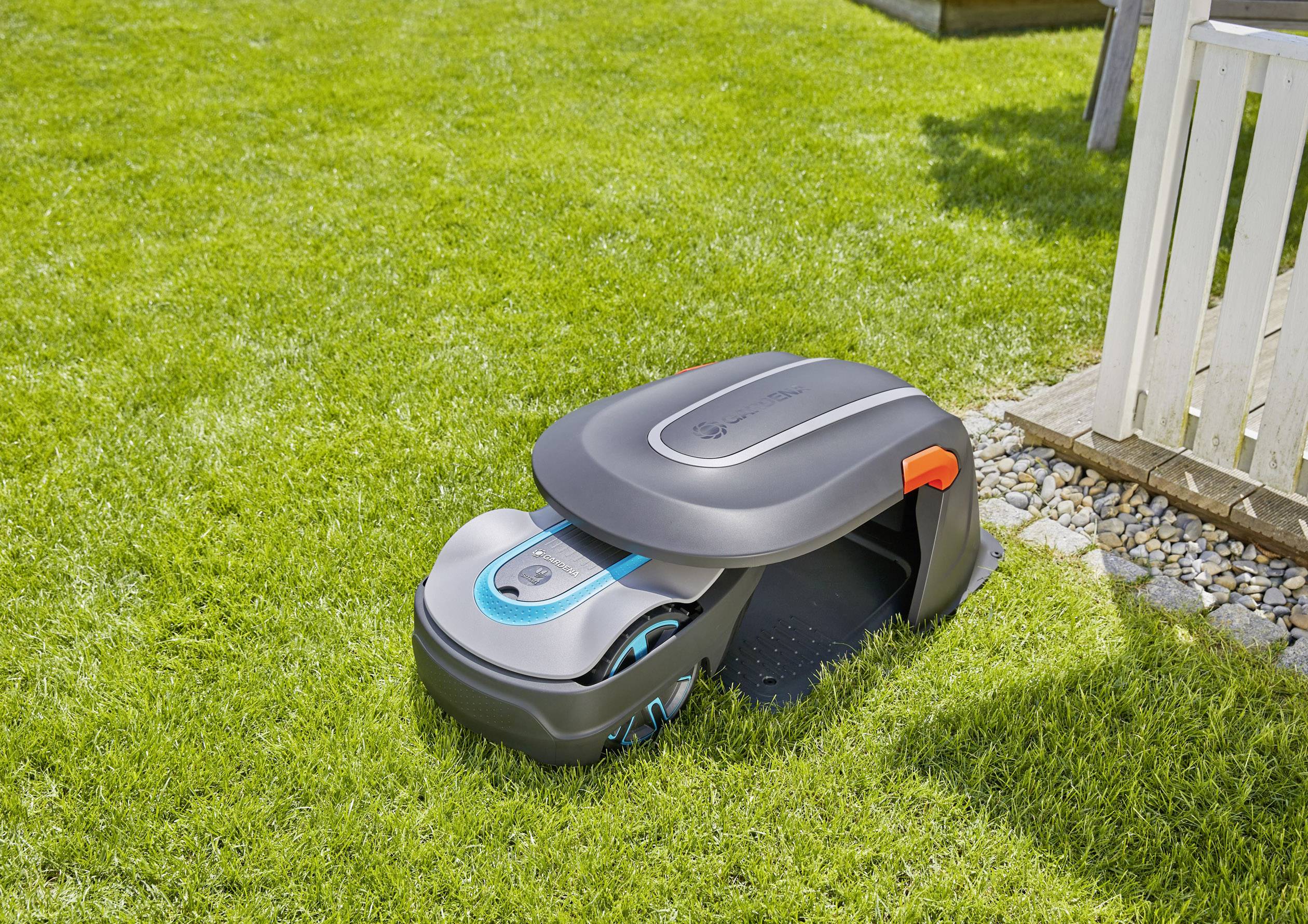 Robot koszący trawę parkuje w stacji ładowania na starannie utrzymanym zielonym trawniku, obok białego płotu.