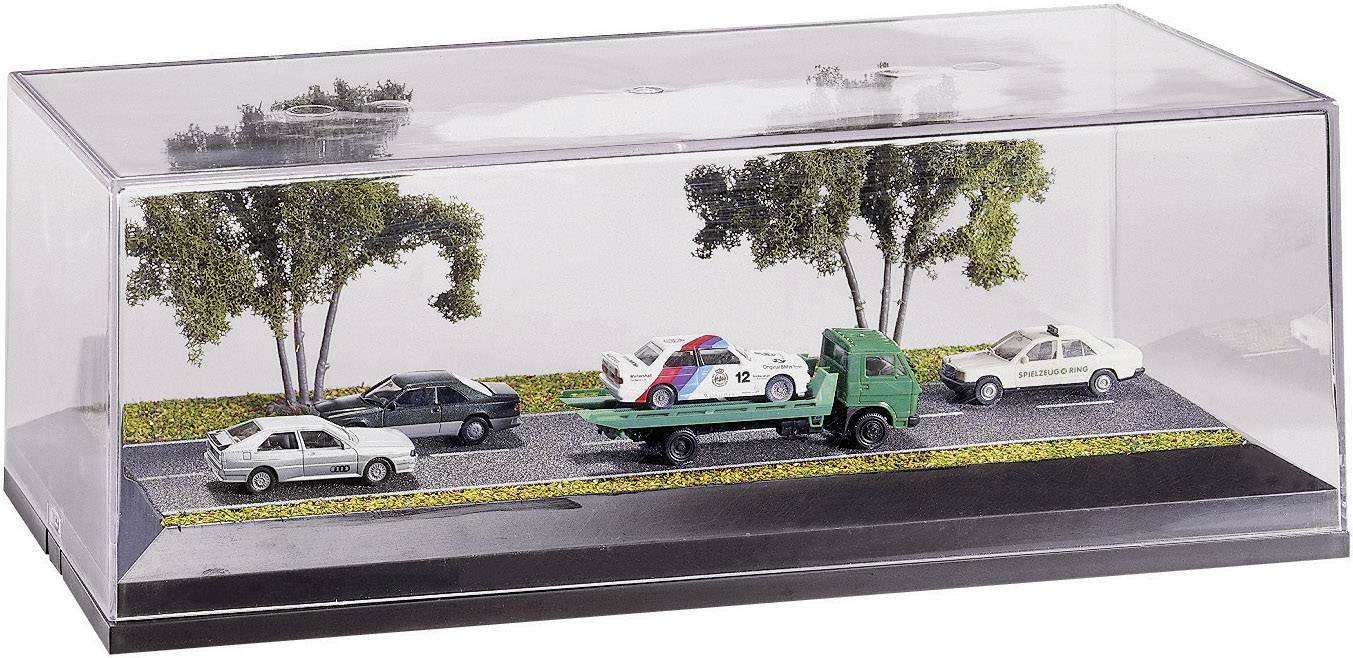 Miniaturowa scena przedstawia pojazd holowniczy transportujący uszkodzony, kolorowy samochód, obok którego znajdują się inne pojazdy i drzewa.