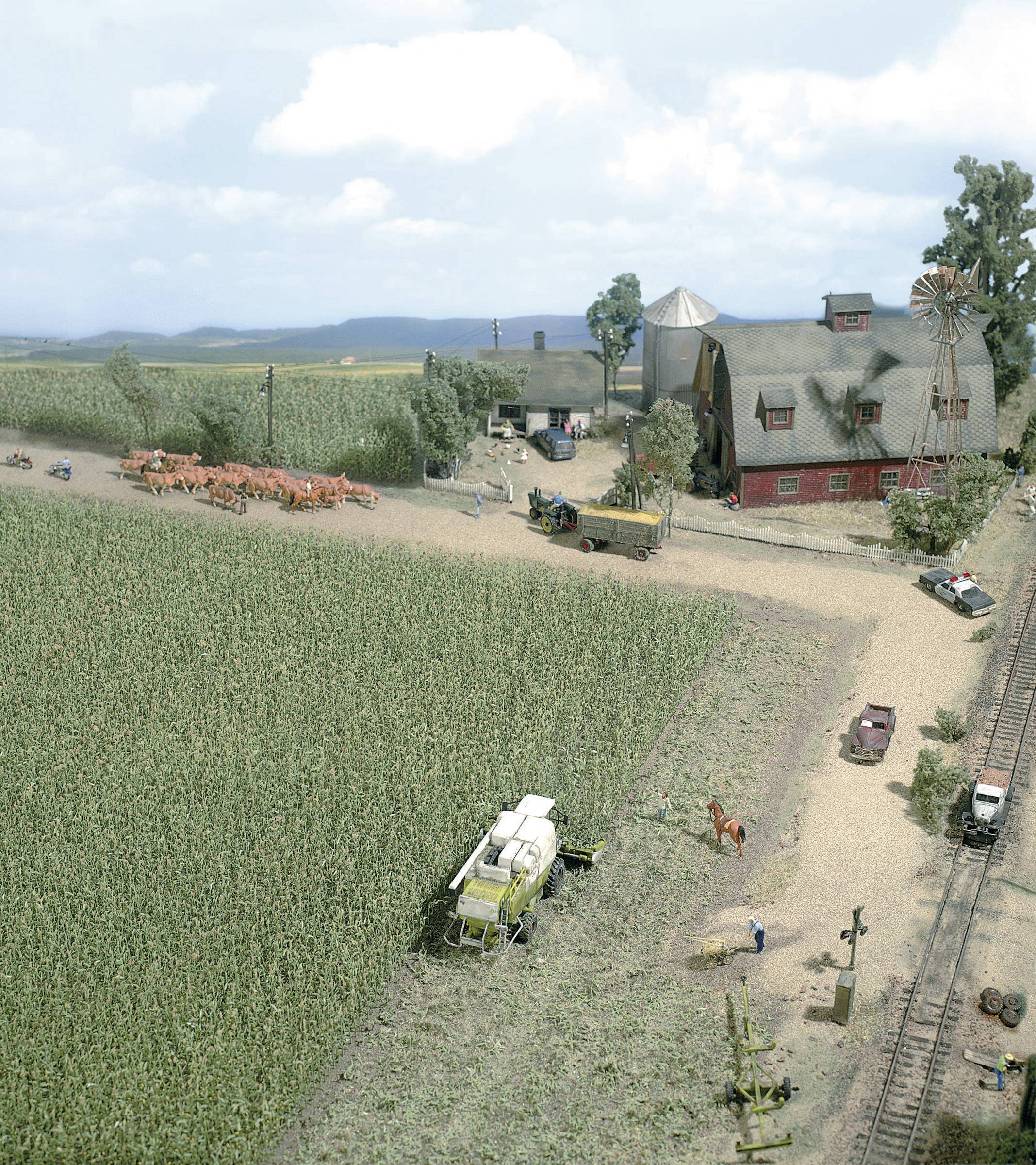 Miniaturowy krajobraz gospodarstwa rolnego z czerwonym budynkiem, krowami na drodze polnej, kombajnem i pojazdami rolniczymi.