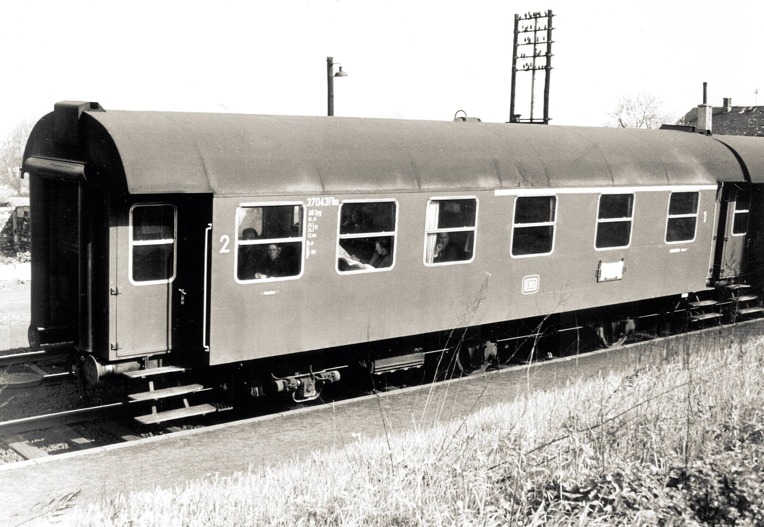 Historyczny wagon kolejowy stoi na peronie. Przez okna widać kilku pasażerów.