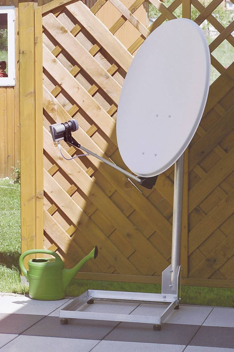 Antena satelitarna skierowana na podwórku obok zielonej konewki i drewnianego płotu, w świetle dziennym.