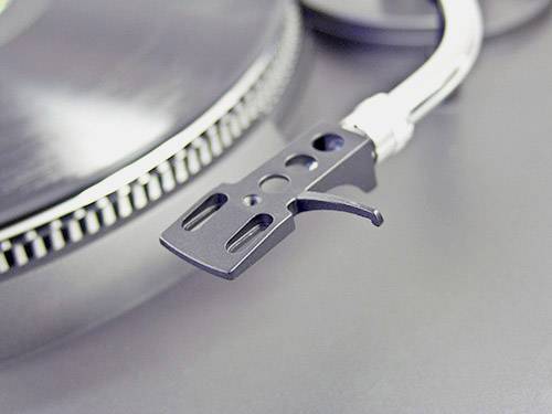 Zbliżenie ramienia gramofonu bez igły, na rozmytym tle talerza płytowego.