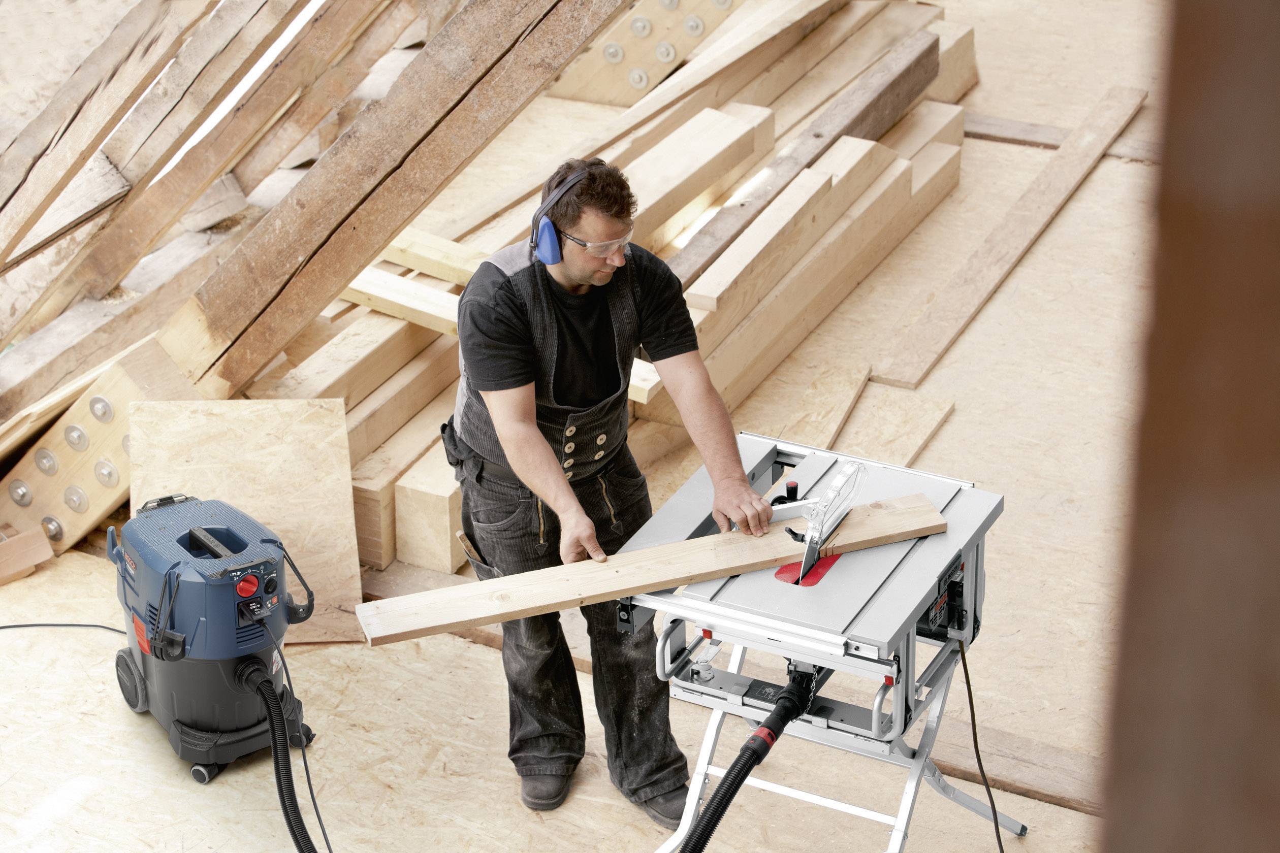 Mężczyzna piłuje deskę drewnianą na placu budowy, używając ochronników słuchu i okularów ochronnych. Posługuje się stołową piłą tarczową, aby wykonać precyzyjne cięcia.