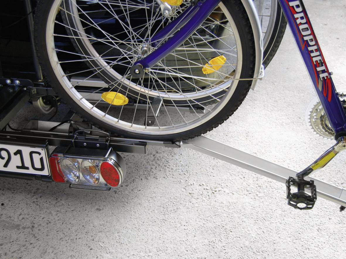 Rower zamocowany na bagażniku samochodu, widoczne przednie koło, częściowo widoczna tablica rejestracyjna, użytkownik w podróży.