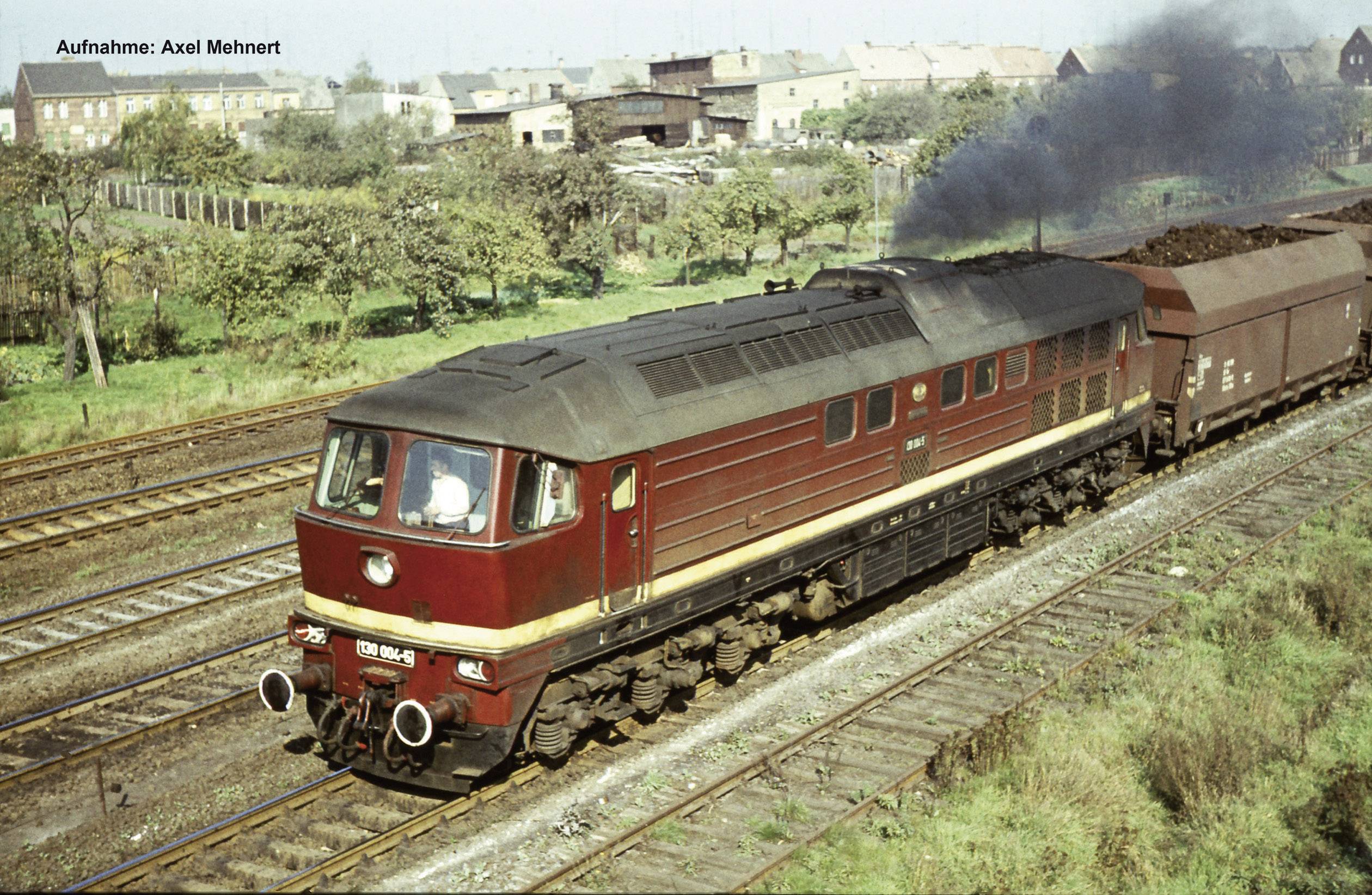 Lokomotywa spalinowa Deutsche Reichsbahn ciągnie pociąg towarowy po torach, z parą i budynkami w tle.