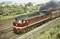 Lokomotywa spalinowa Deutsche Reichsbahn ciągnie pociąg towarowy po torach, z parą i budynkami w tle.