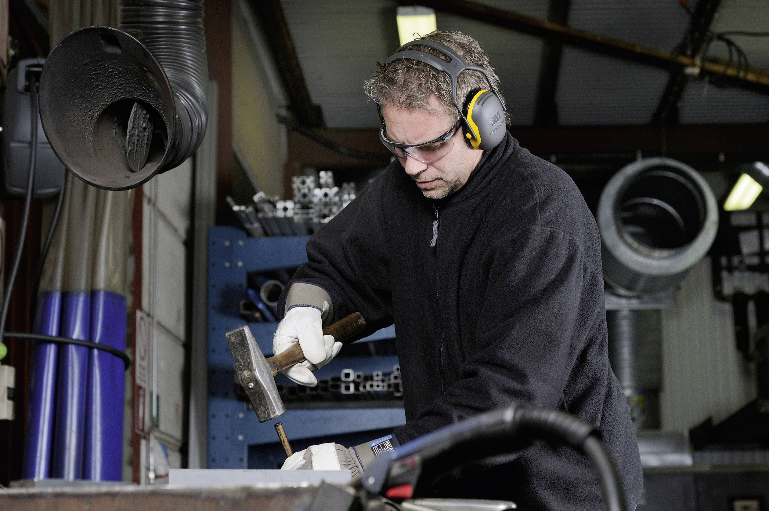 Mężczyzna w odzieży ochronnej i ochronnikach słuchu pracuje w warsztacie, uderzając młotkiem w kawałek metalu. W tle widoczne są regały z materiałami.