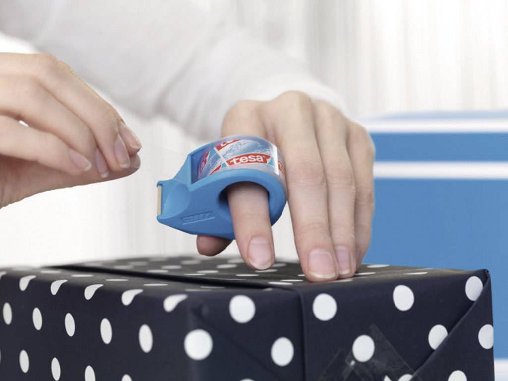 Jedna ręka używa niebieskiego dozownika do palców, aby zapakować prezent w papier w kropki.