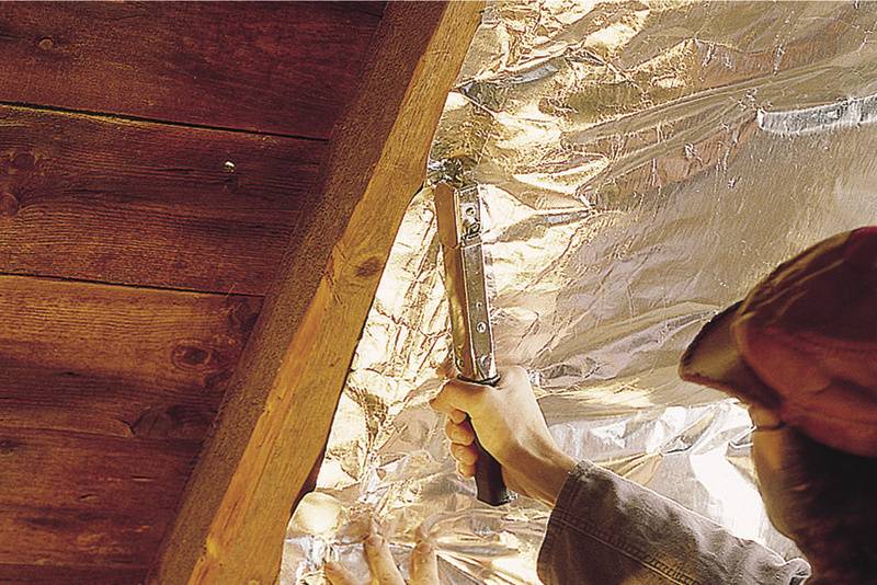 Osoba mocuje izolacyjną folię do więźby dachowej za pomocą zszywacza. Folia odbija światło.