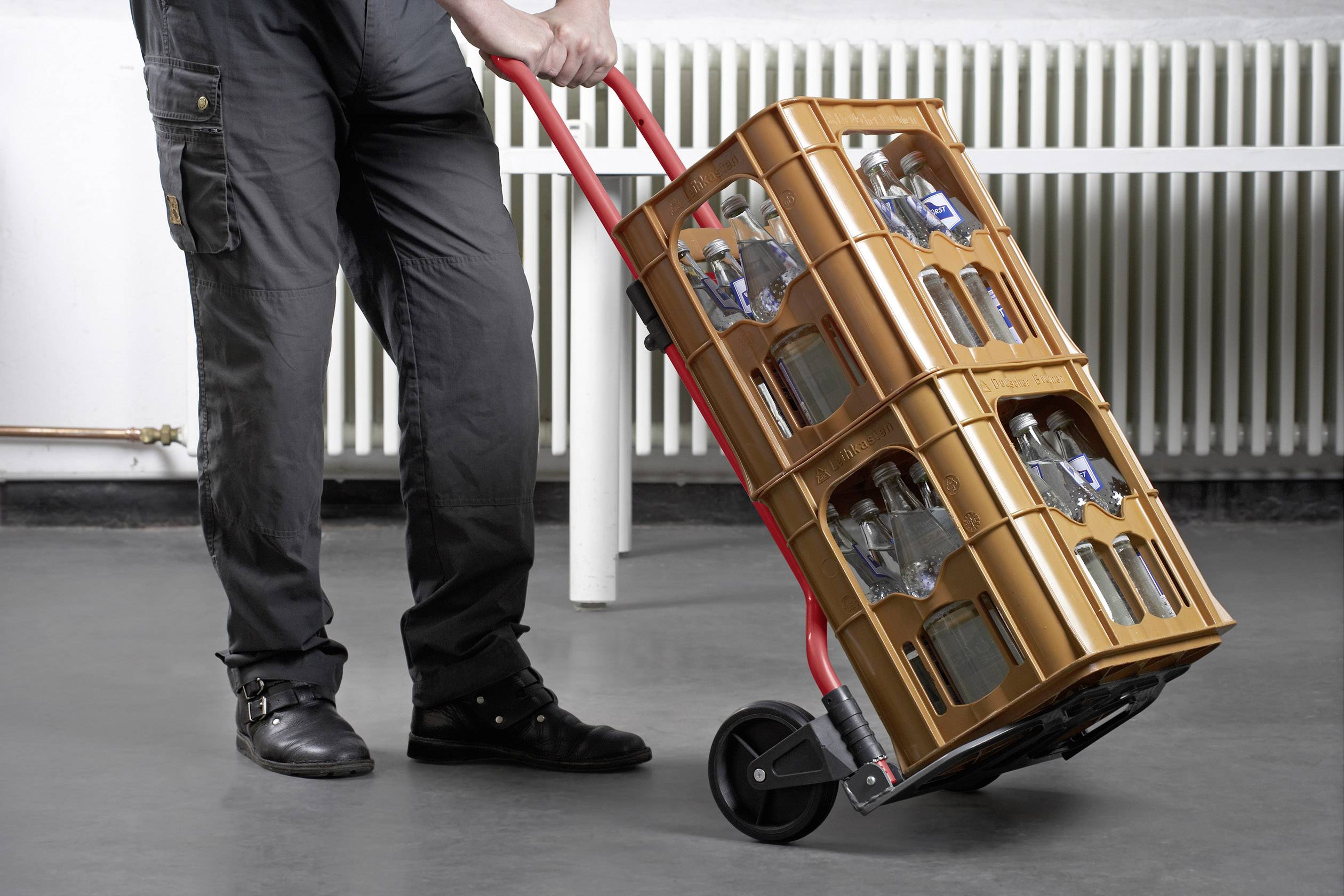 Osoba przesuwa taczkę z dwoma skrzynkami pustych butelek szklanych w środowisku przemysłowym.