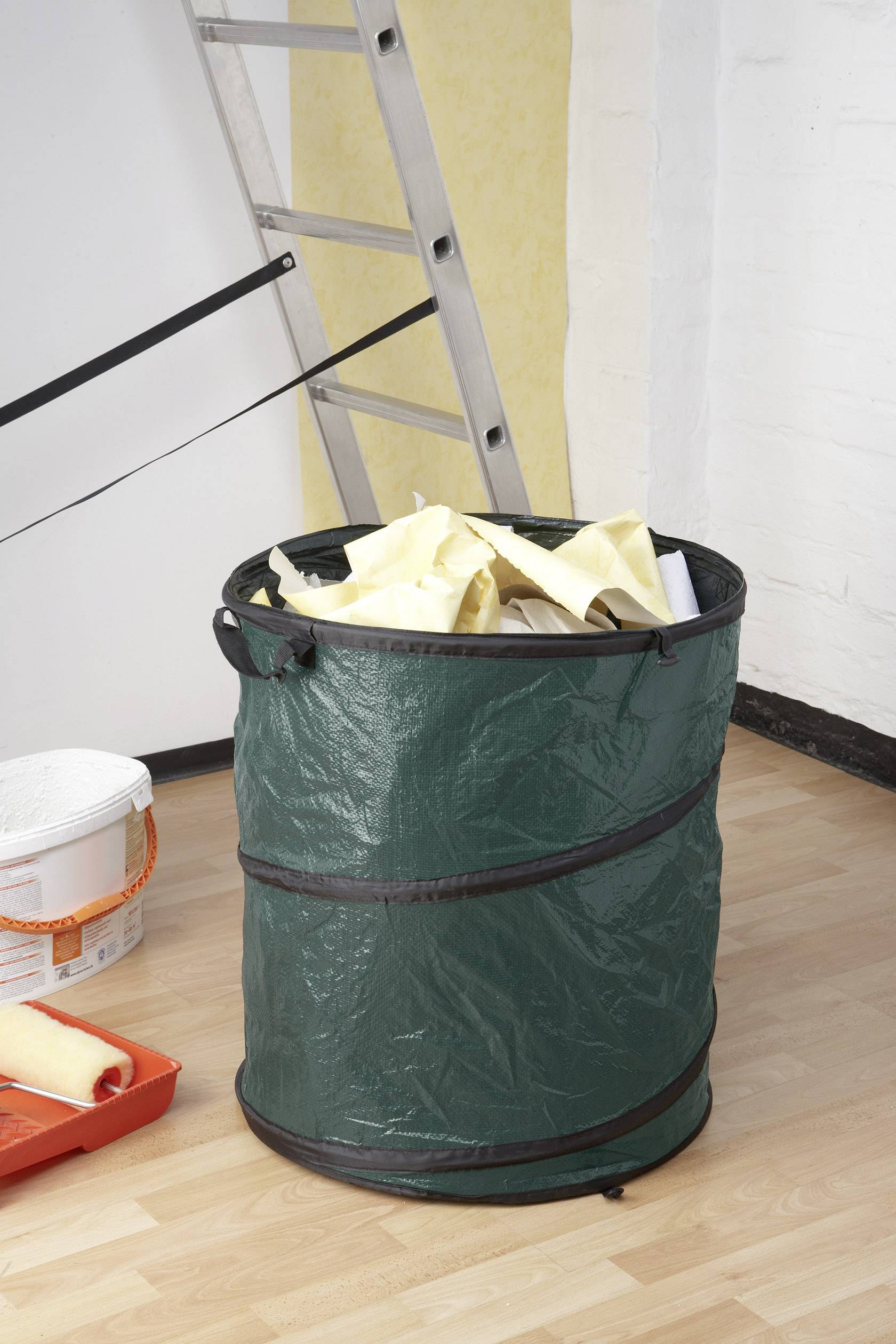 Duży zielony worek na śmieci, wypełniony resztkami tapet, stoi na drewnianej podłodze obok drabiny i wiader z farbą.