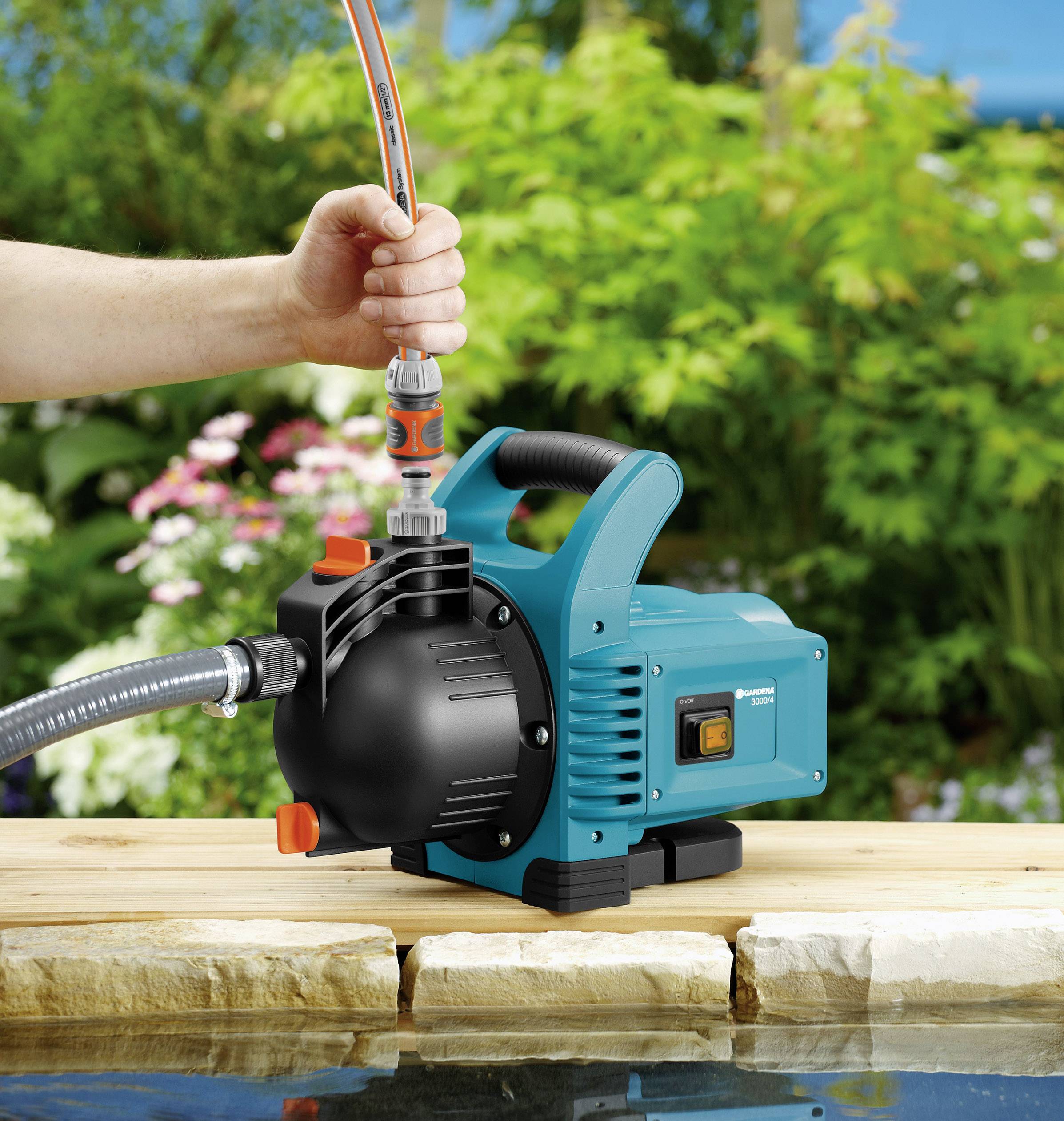 Osoba instaluje niebieską pompę ogrodową obok stawu, otoczoną roślinami. Pompa jest gotowa do tłoczenia wody.