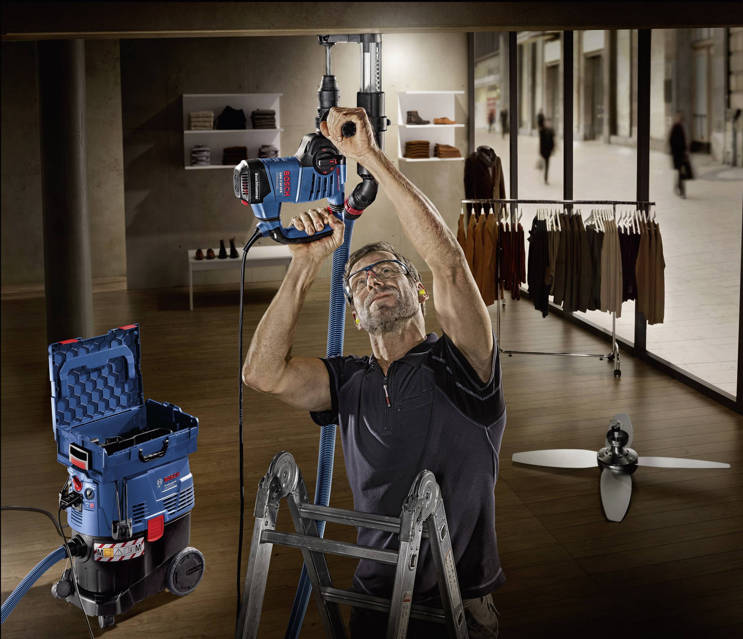 Mężczyzna stoi na drabinie w sklepie odzieżowym i wierca w suficie elektronarzędziem. Na dole stoi odkurzacz.