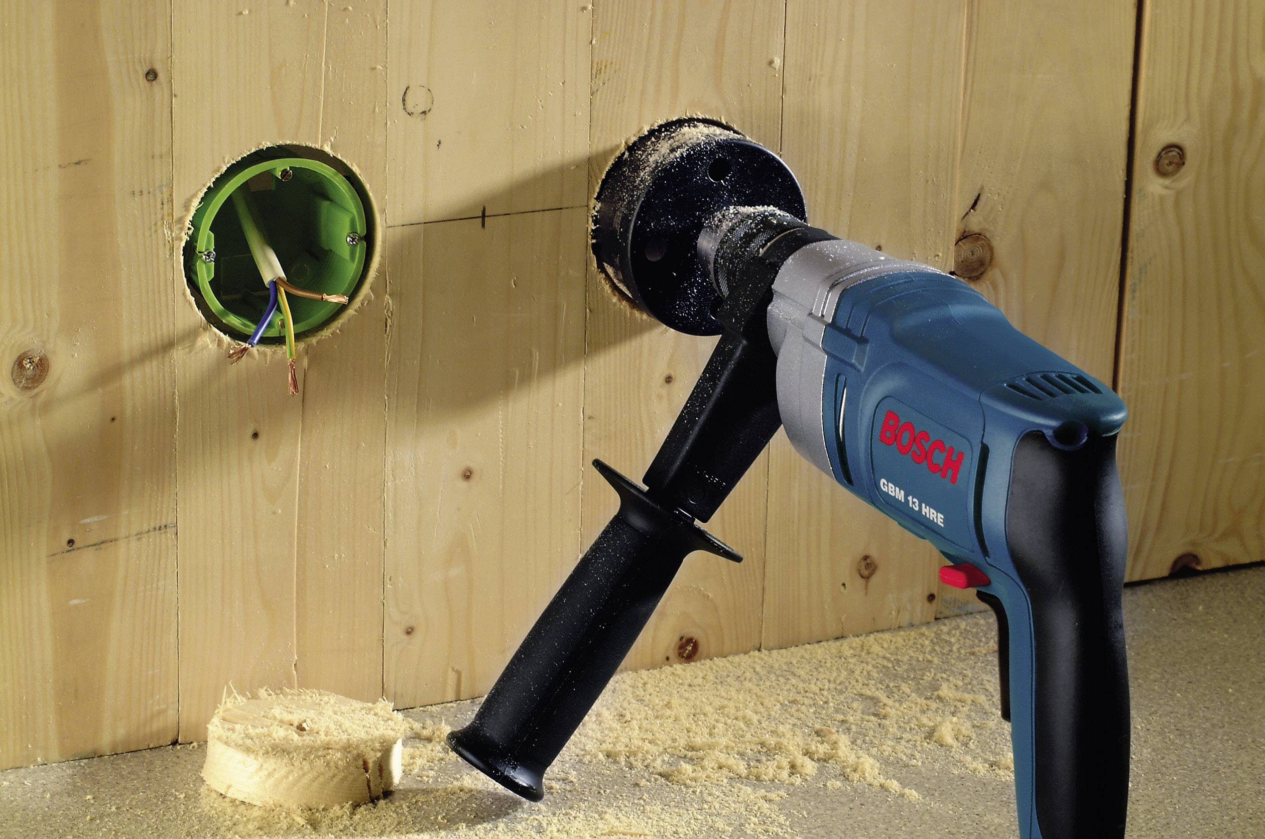 Wiertarka z piłą otworową wywierca okrągły otwór w płycie drewnianej obok elektrycznej puszki przyłączeniowej.