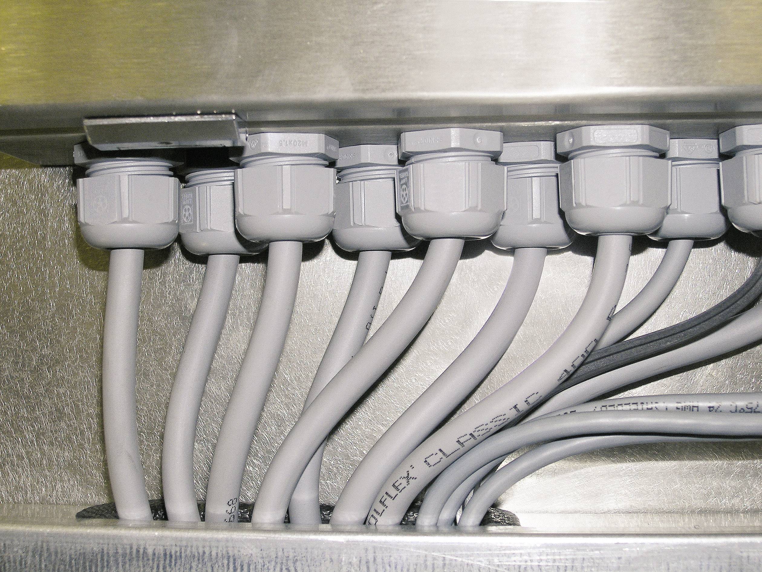 Kilka szarych przewodów biegnie przez dławnice kablowe w metalowej skrzynce, ułożonych obok siebie w sposób uporządkowany.