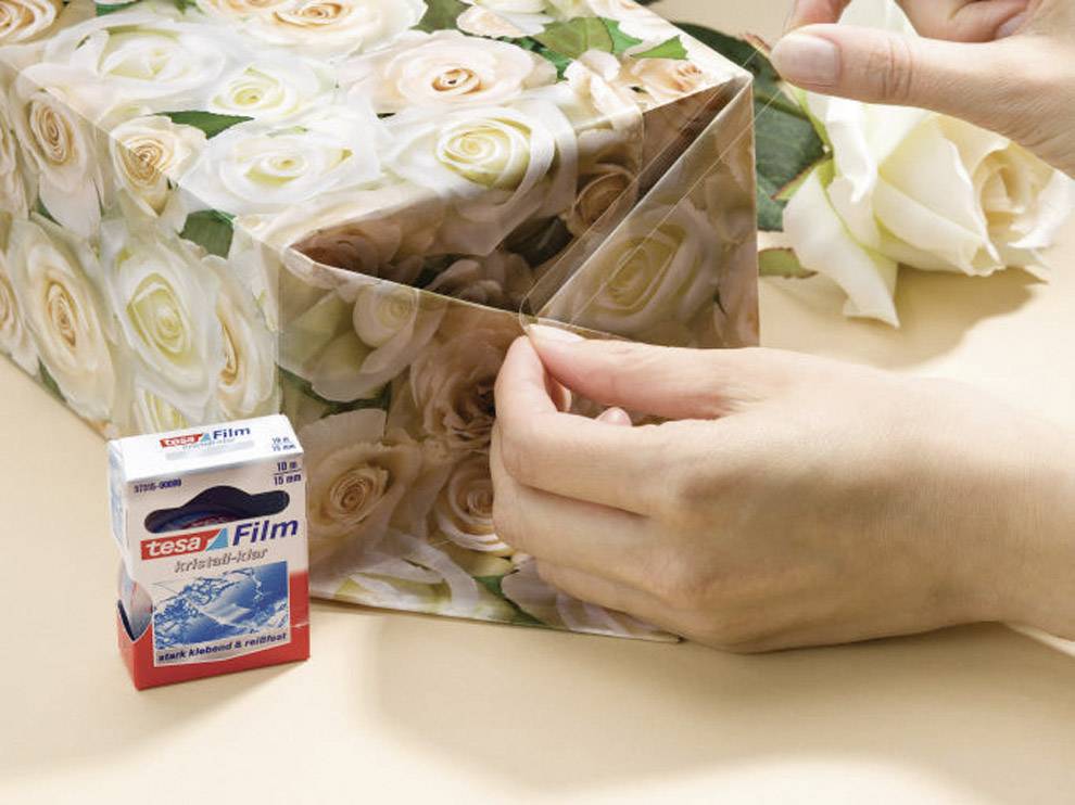 Osoba pakuje prezent w papier w róże i używa taśmy klejącej. Obok ręki leży rolka taśmy Tesa.