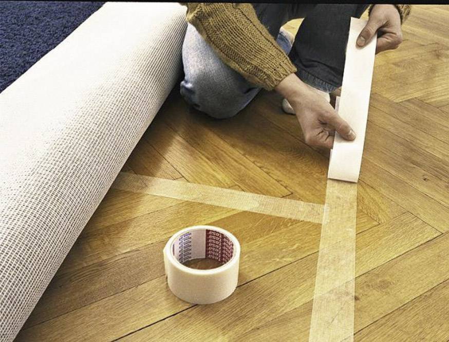 Ktoś nakłada taśmę dwustronną na drewnianą podłogę, aby ułożyć dywan. W tle leży zwinięty dywan.