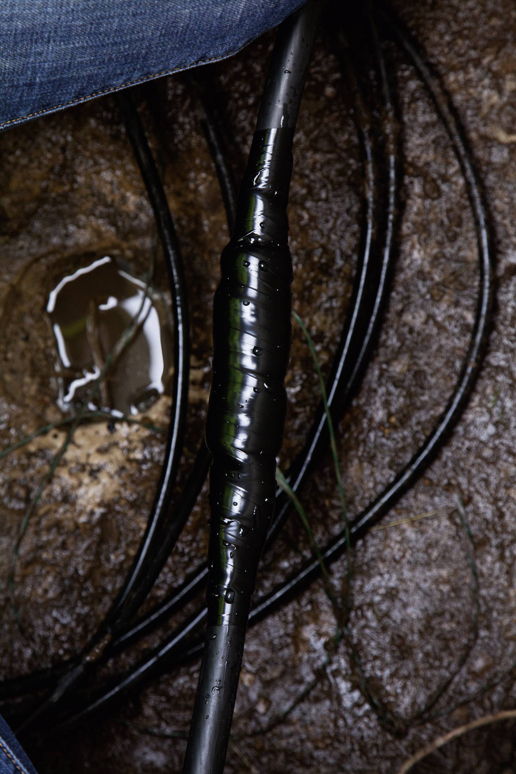 Czarny kabel owinięty taśmą izolacyjną leży na mokrej ziemi. Krople wody widoczne są na taśmie, otoczone ziemią.