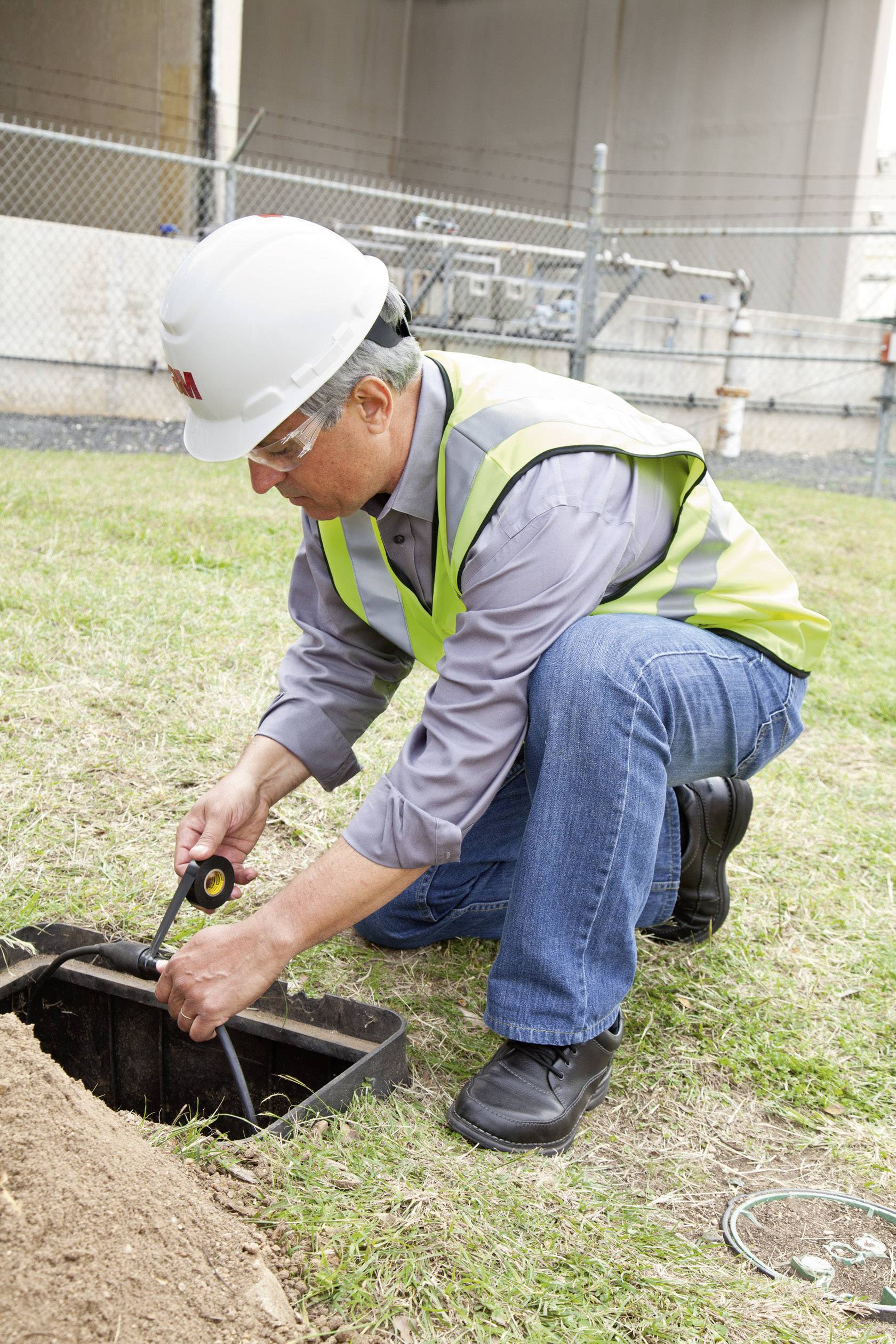 Robotnik w kasku ochronnym i kamizelce ostrzegawczej sprawdza skrzynkę kablową w ziemi. W tle widoczny jest płot.