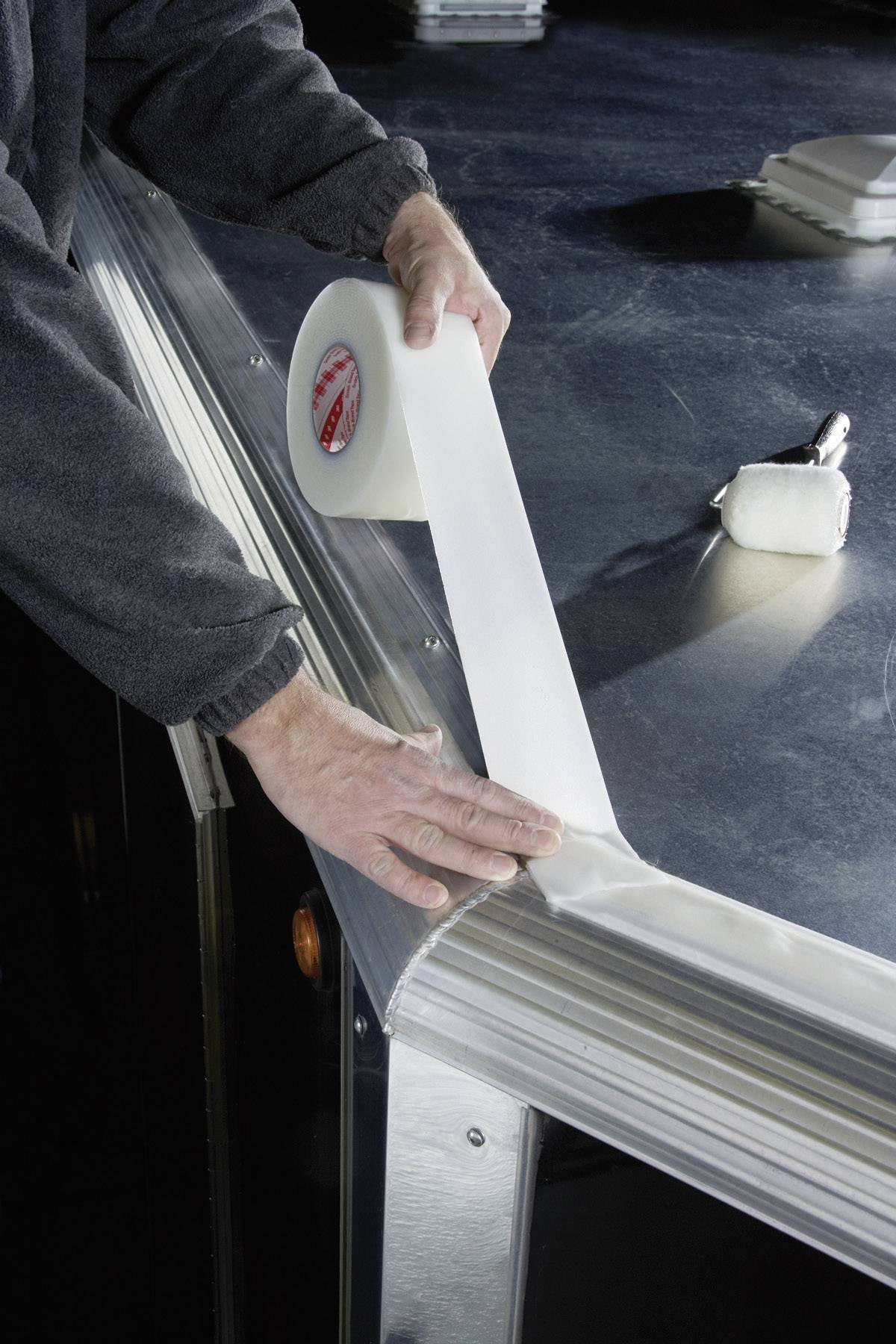 Osoba nakłada rolkę przezroczystej taśmy klejącej na krawędź metalowej powierzchni, prawdopodobnie w celu ochrony lub uszczelnienia.