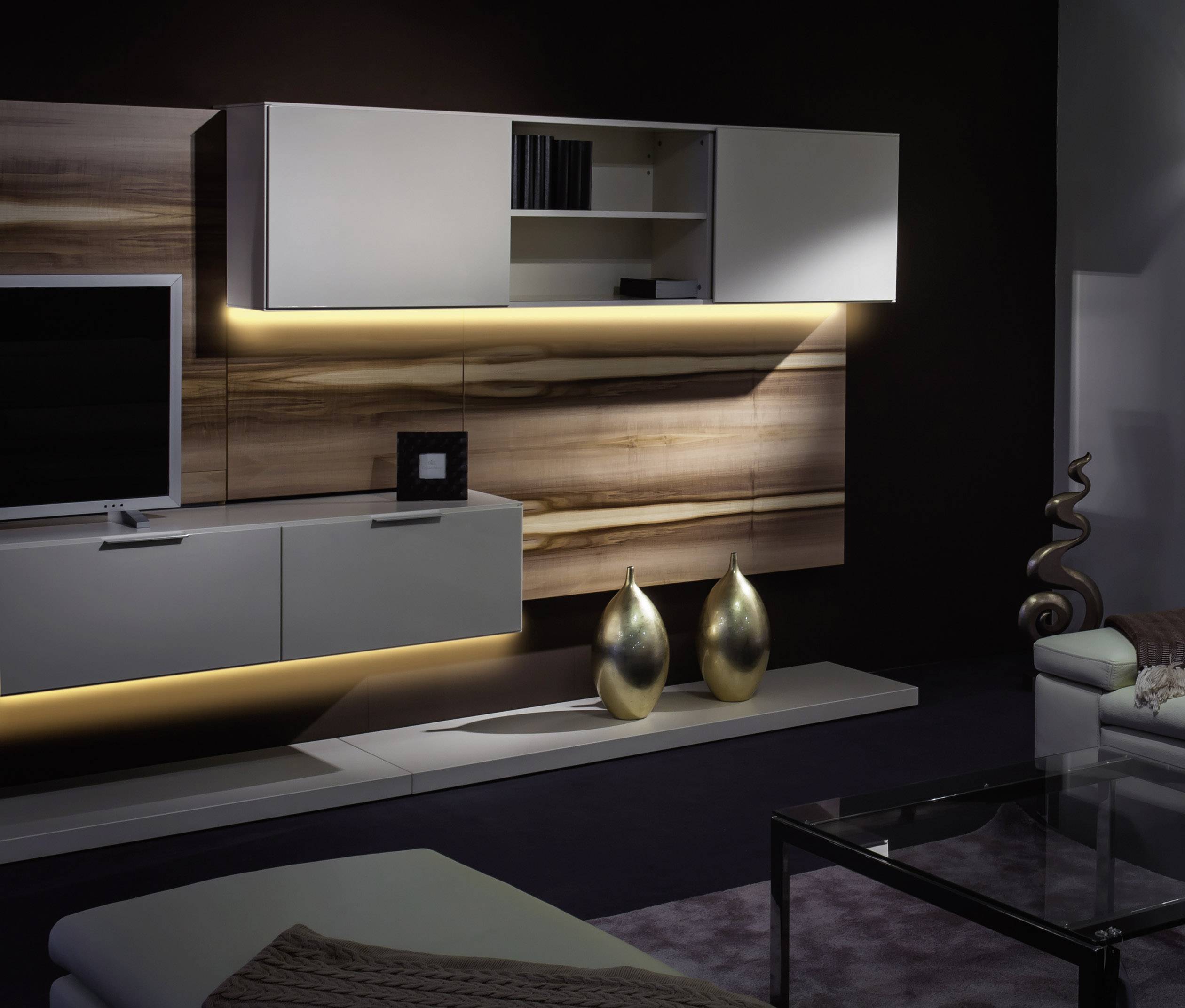 Salon z nowoczesną, podświetlaną szafką ścienną w wyglądzie drewna, telewizorem, szklanym wazonem, elementami dekoracyjnymi i elegancką sofą.