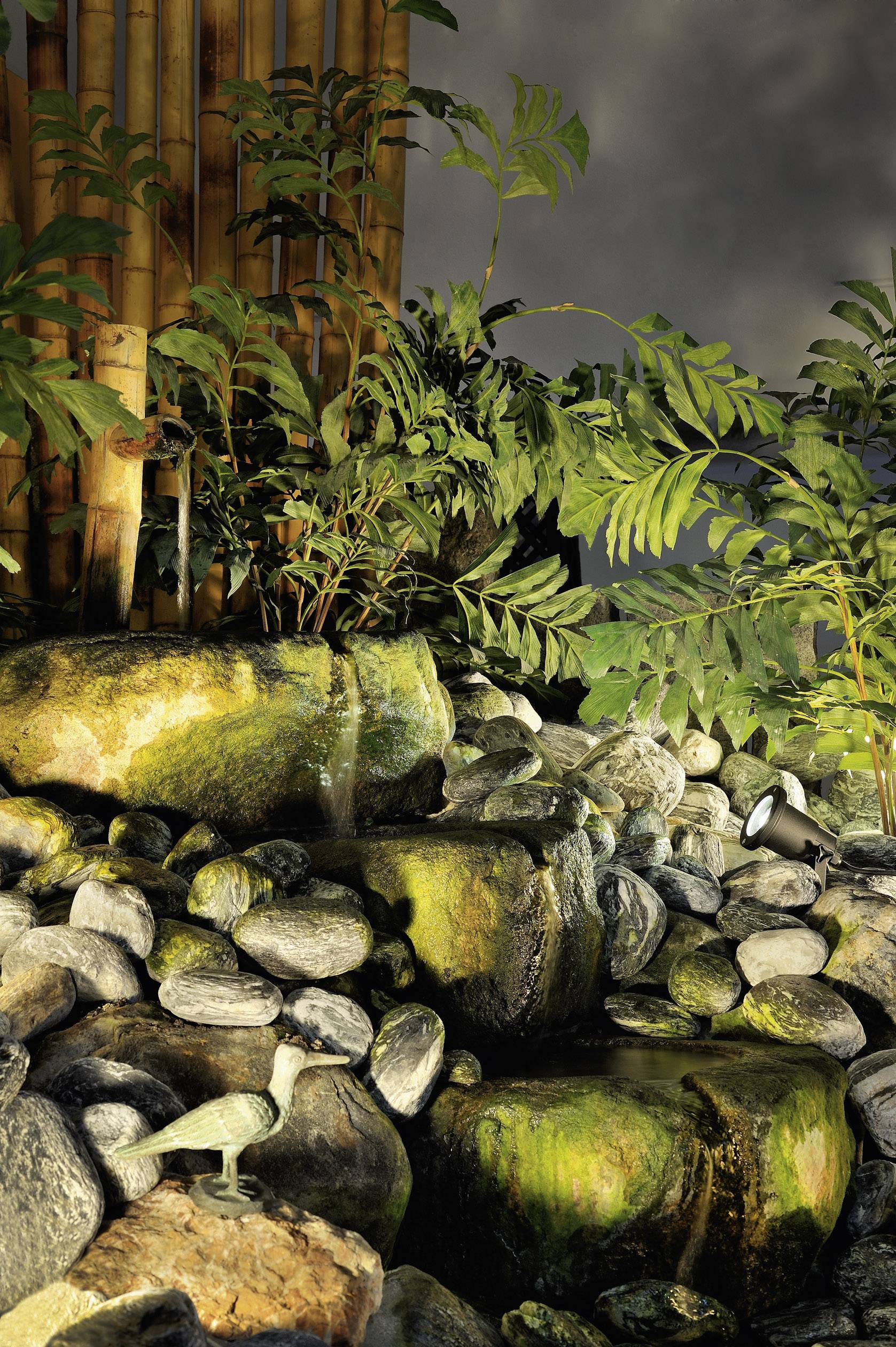 Mały, skalisty wodospad w ogrodzie otoczonym roślinnością. W tle widoczne są bambusowe kije.