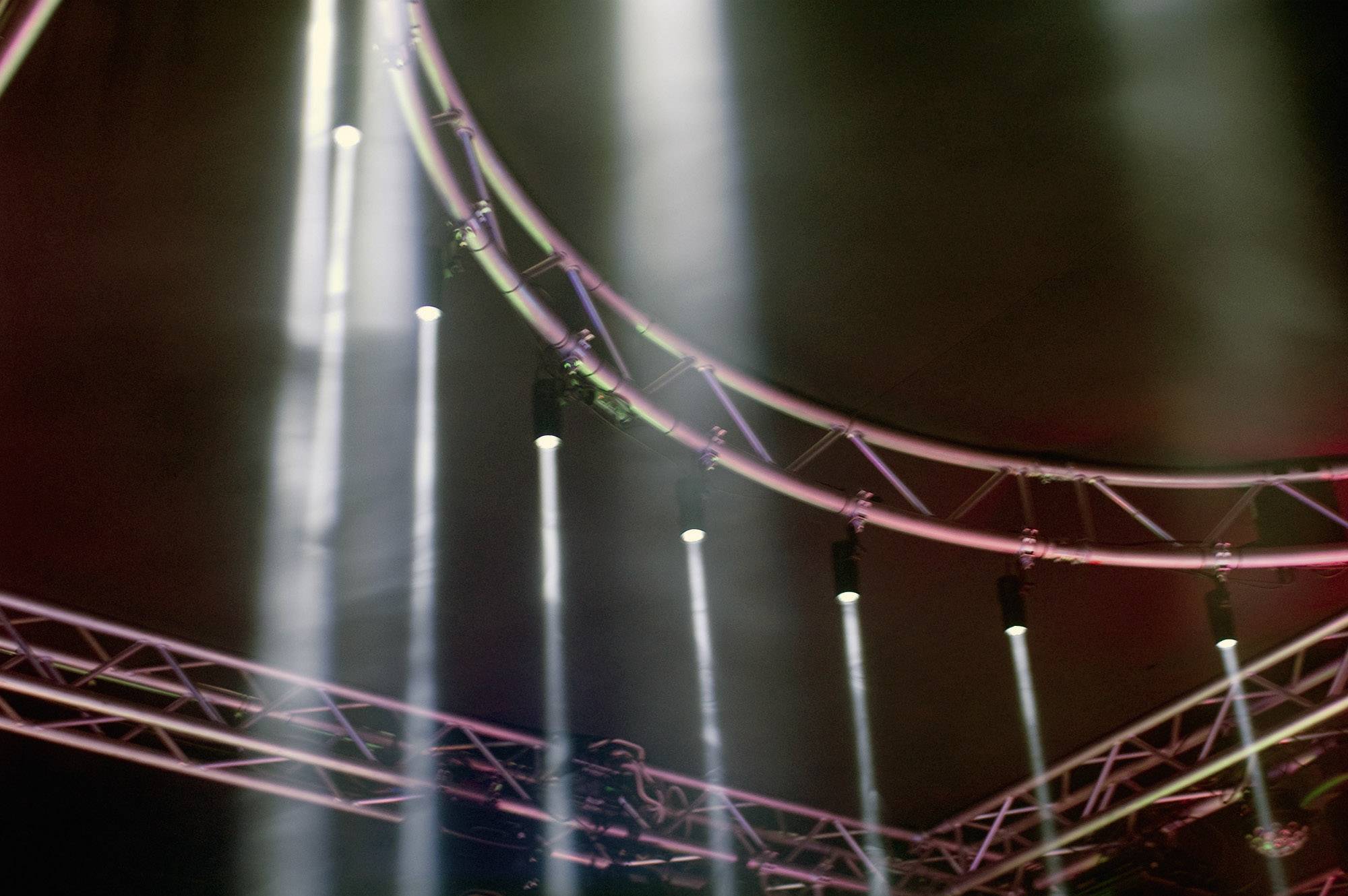 Oświetlenie sceniczne z góry, z wieloma białymi reflektorami skierowanymi w dół. Widoczne są struktury nośne, ciemne tło.