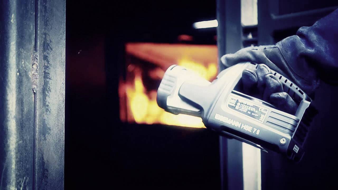 Osoba trzyma termometr na podczerwień przed otwartym piecem z widocznymi płomieniami, mierząc jego temperaturę.