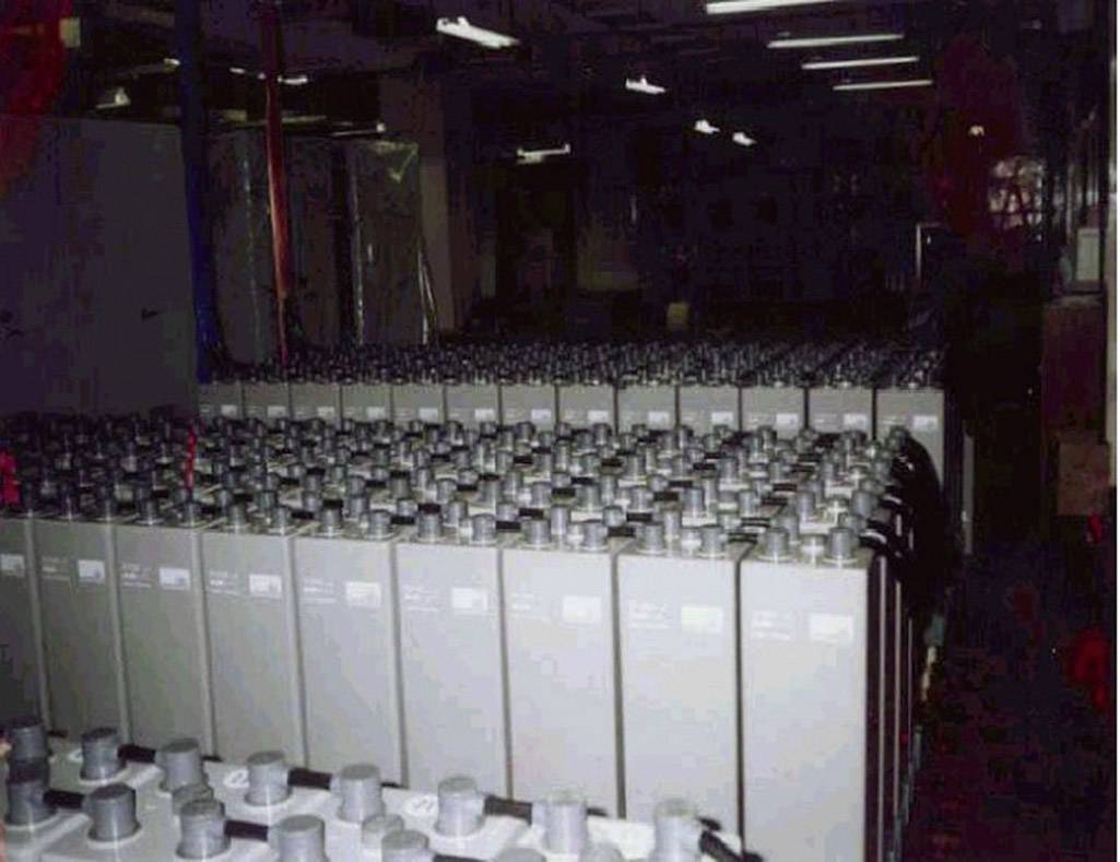 Rząd dużych szarych baterii stoi w magazynie, ułożony w równych rzędach. Oświetlenie jest słabe.
