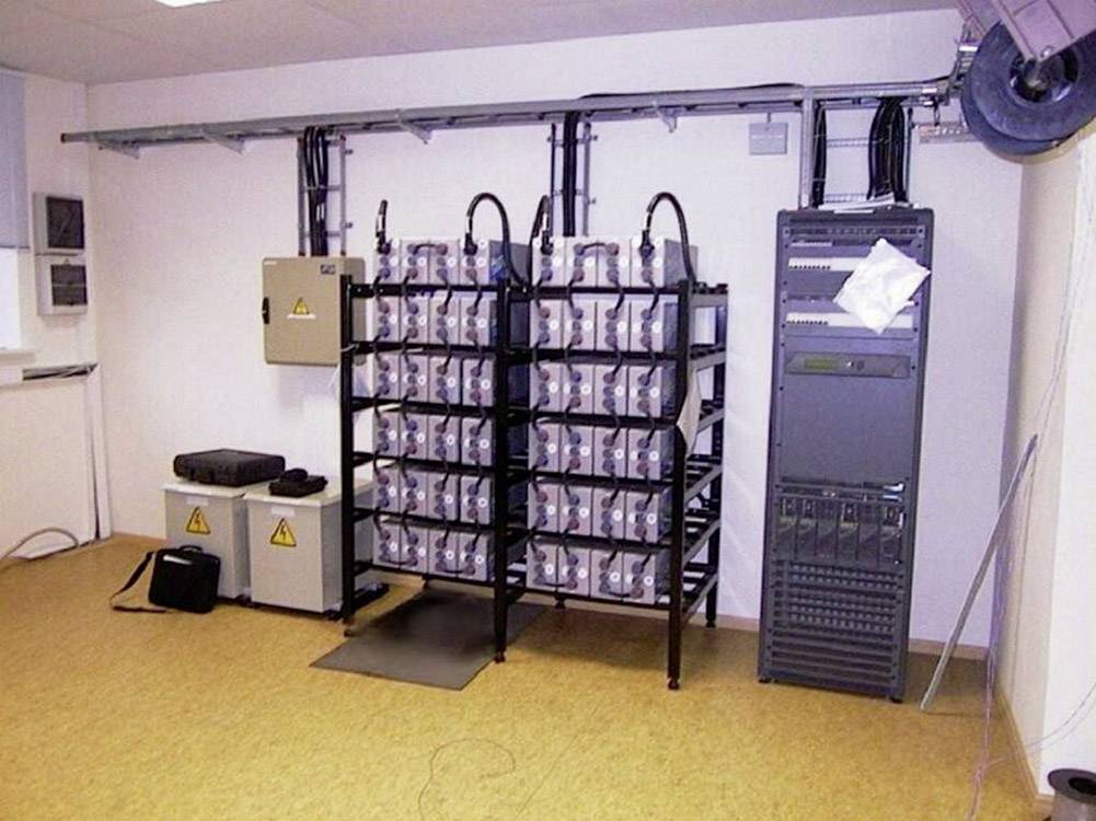Serwerownia z wieloma zasilaczami UPS, skrzynką zabezpieczeń i szafą serwerową. Okablowanie przymocowane do ściany.