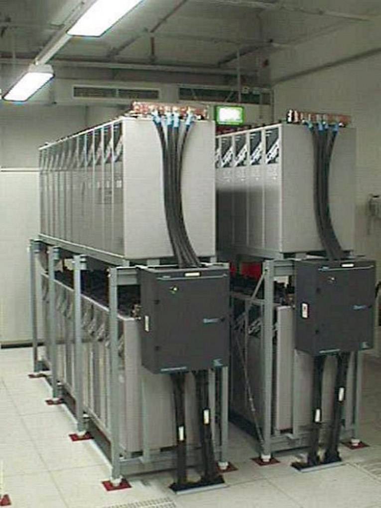 Centrum obliczeniowe z dwoma rzędami dużych, szarych szaf serwerowych z kablami prowadzonymi u góry, w białym, dobrze oświetlonym pomieszczeniu.