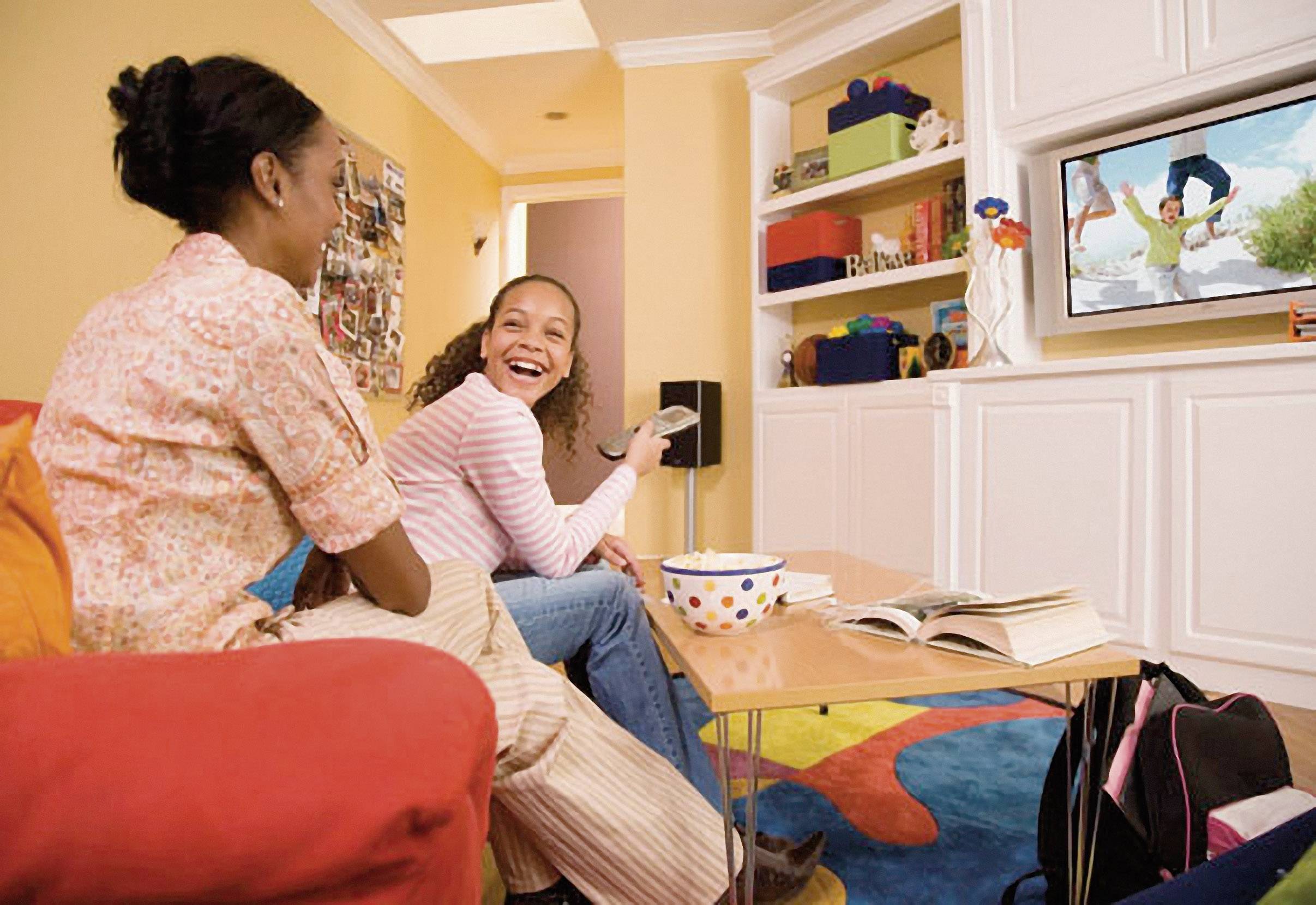 Dwie kobiety siedzą na kanapie w salonie i śmieją się, oglądając telewizję. Na stole leży otwarta książka i paczka popcornu.