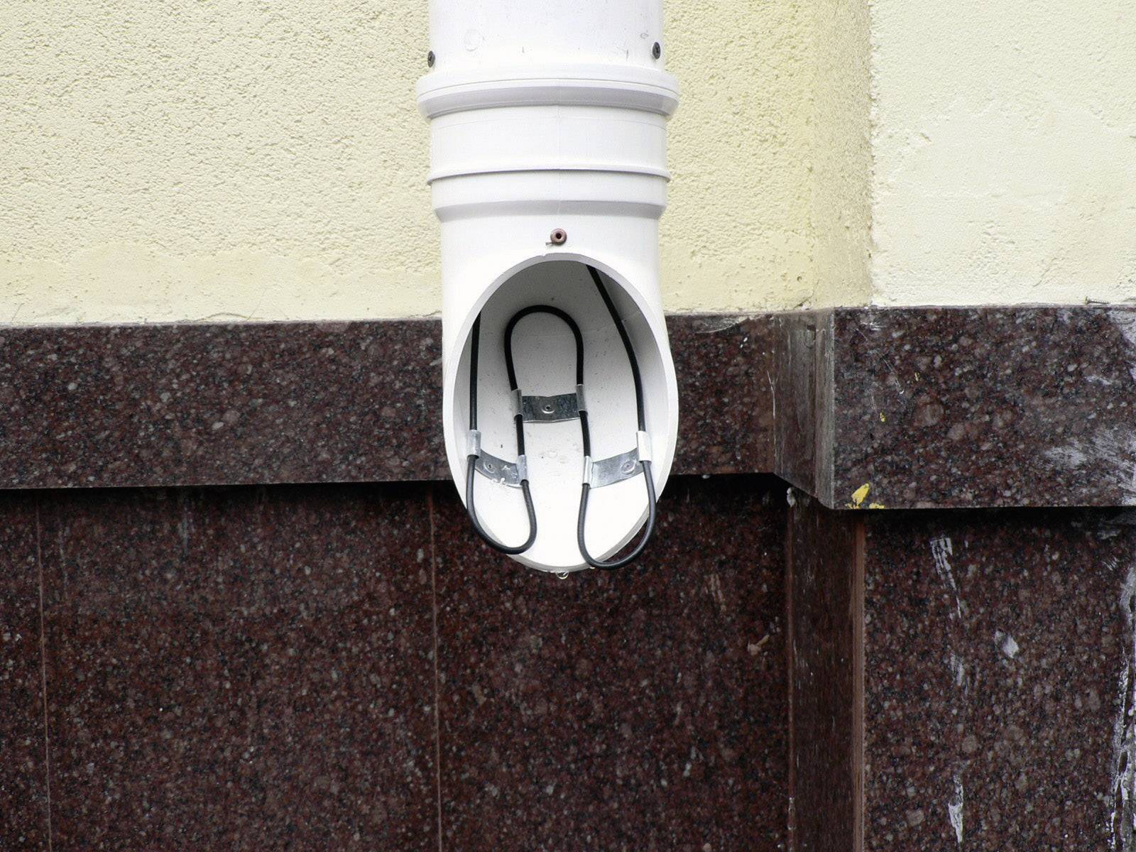 Biała atrapa rury odpływowej na ścianie, przypominająca kamerę monitoringu. Humorystyczna i zwodnicza instalacja.
