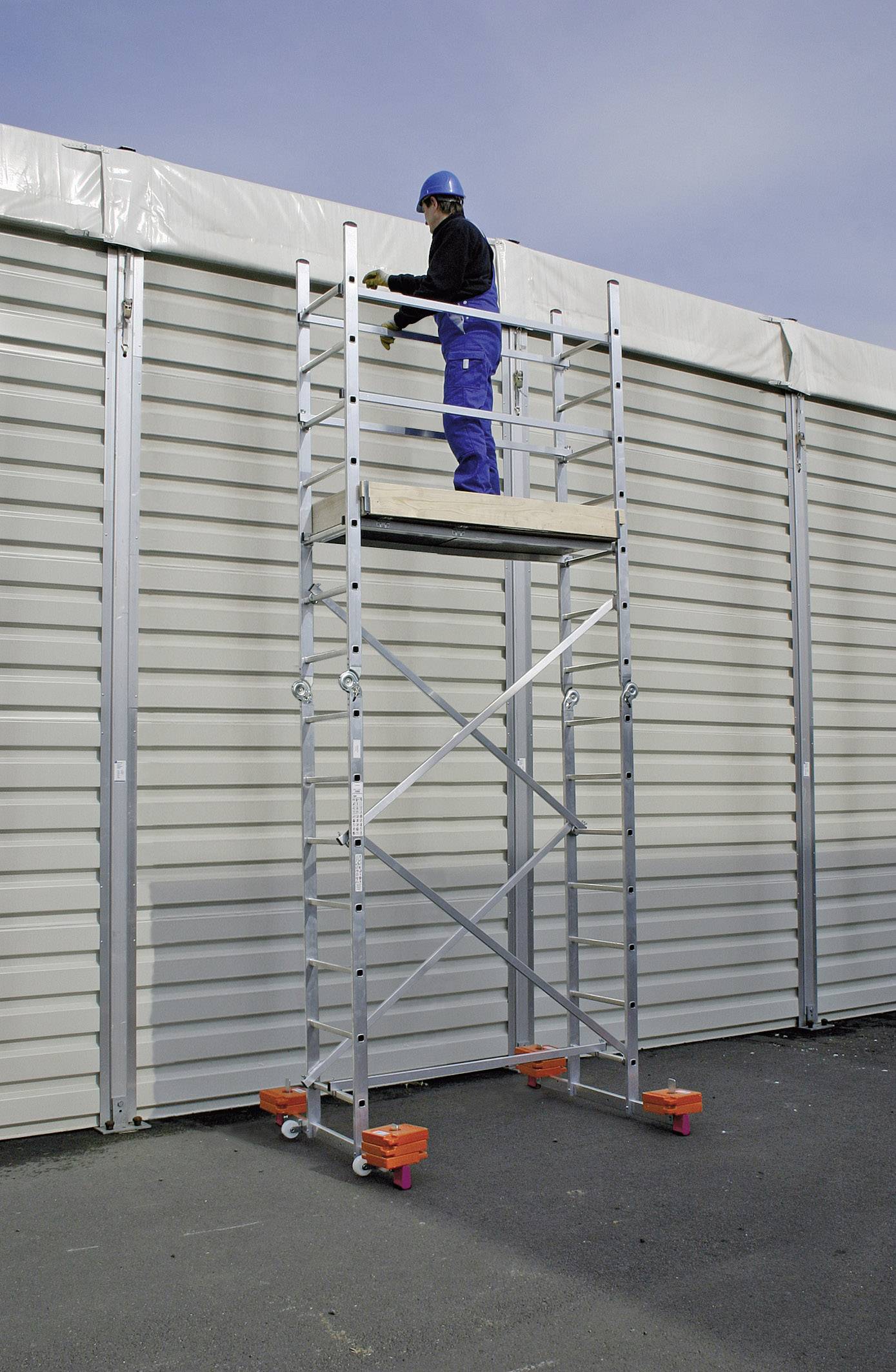 Robotnik stoi na ruchomym rusztowaniu przed dużą metalową ścianą. Jest ubrany w odzież ochronną, w tym kask.