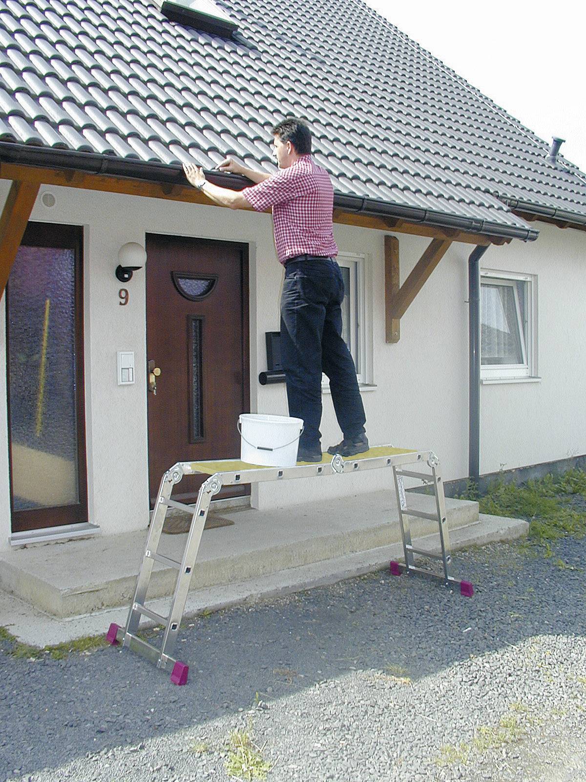 Osoba stoi na drabinie i czyści rynnę domu jednorodzinnego. Wiadro stoi na drabinie.