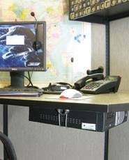 Biurko z komputerem, telefonem i myszą. Na tle ściany wisi mapa świata. Obok monitora leży słuchawka z mikrofonem.