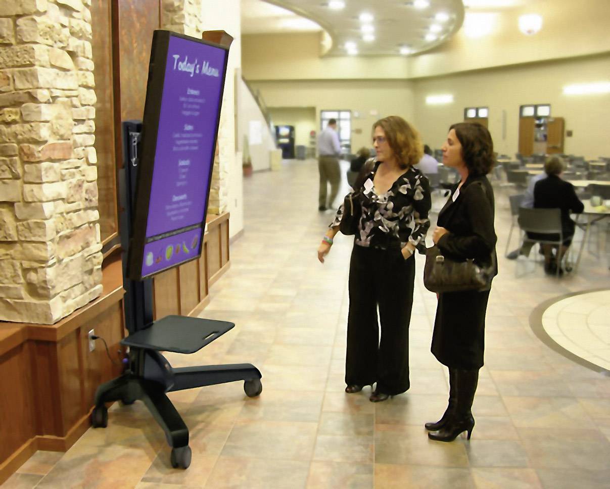 Dwie kobiety przeglądają cyfrowe menu na ekranie w dużej, nowoczesnej hali, w tle widać stoły i krzesła.