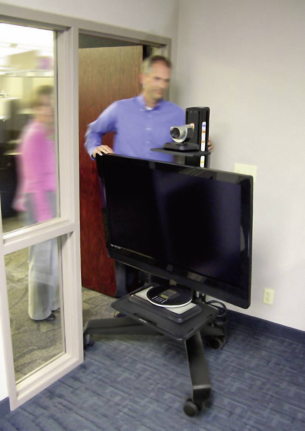 Mężczyzna przesuwa wózek konferencyjny z dużym ekranem i kamerą przez drzwi biurowe. W tle widać drugą osobę.