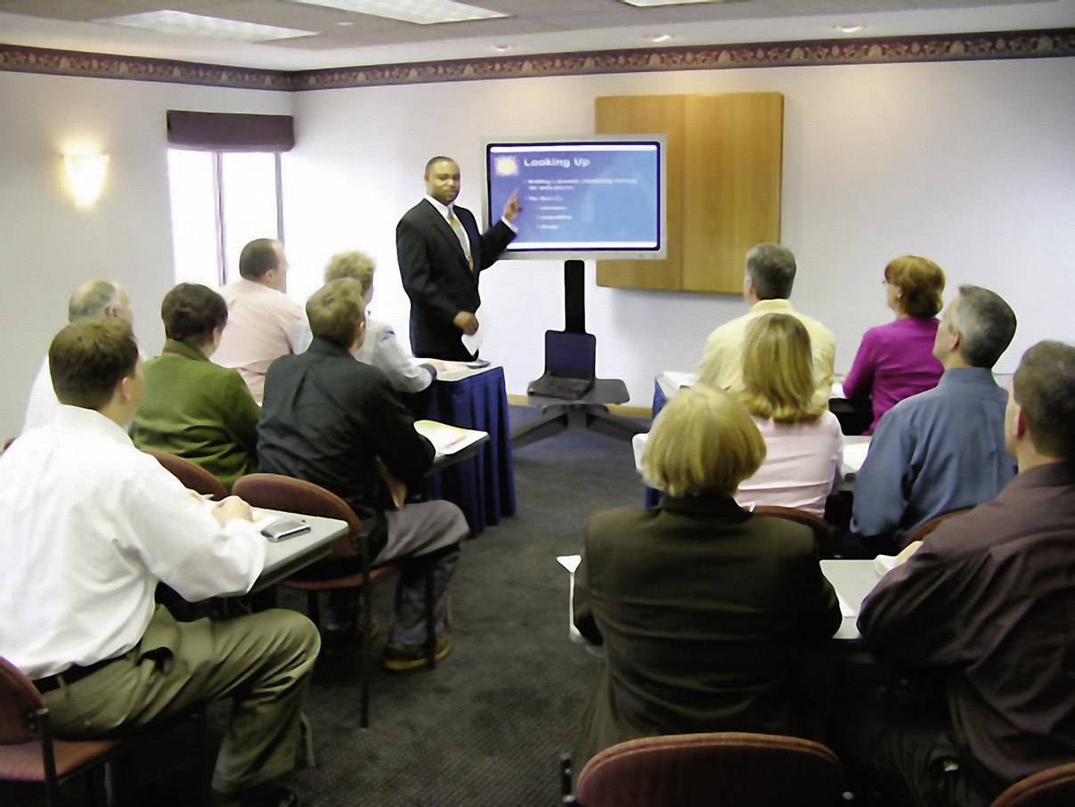 Mężczyzna prowadzi prezentację przed grupą osób siedzących w sali konferencyjnej. Wszyscy są skupieni na ekranie.